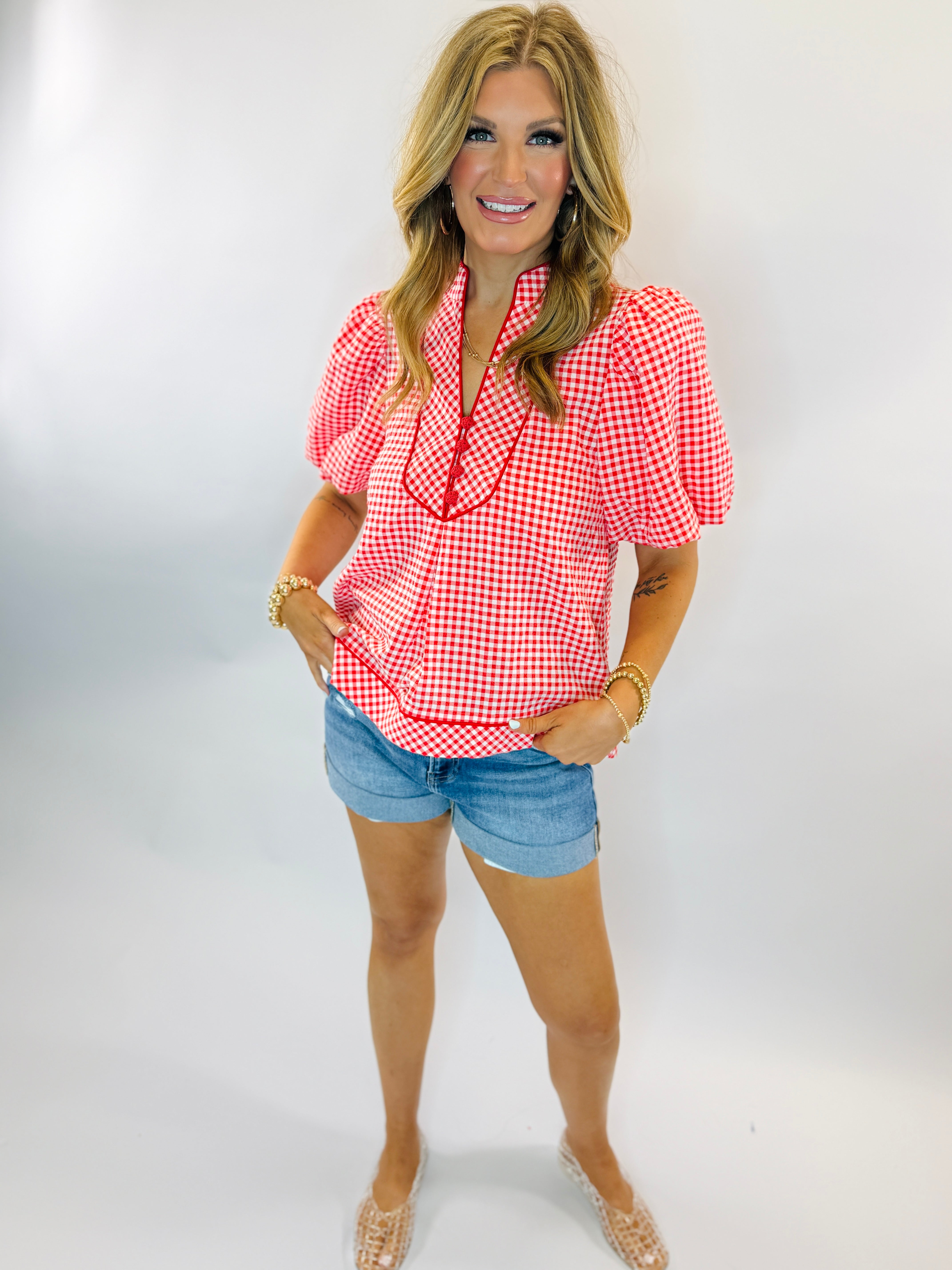 Woman wearing red and white gingham Diana top with bow tie neck and puffed sleeves, paired with denim shorts.