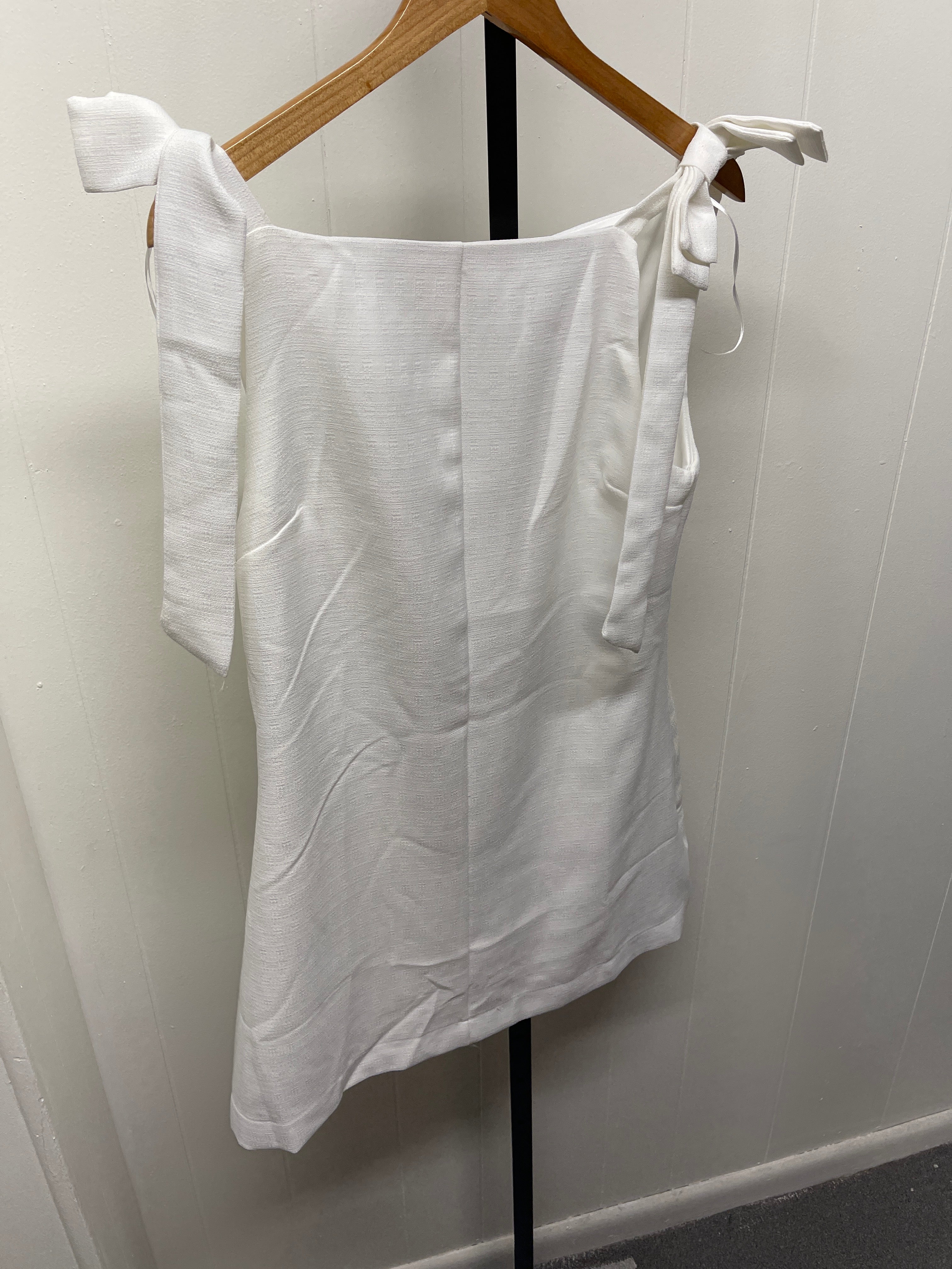 White sleeveless mini dress with tie shoulder straps hanging on wooden hanger against white tiled wall.