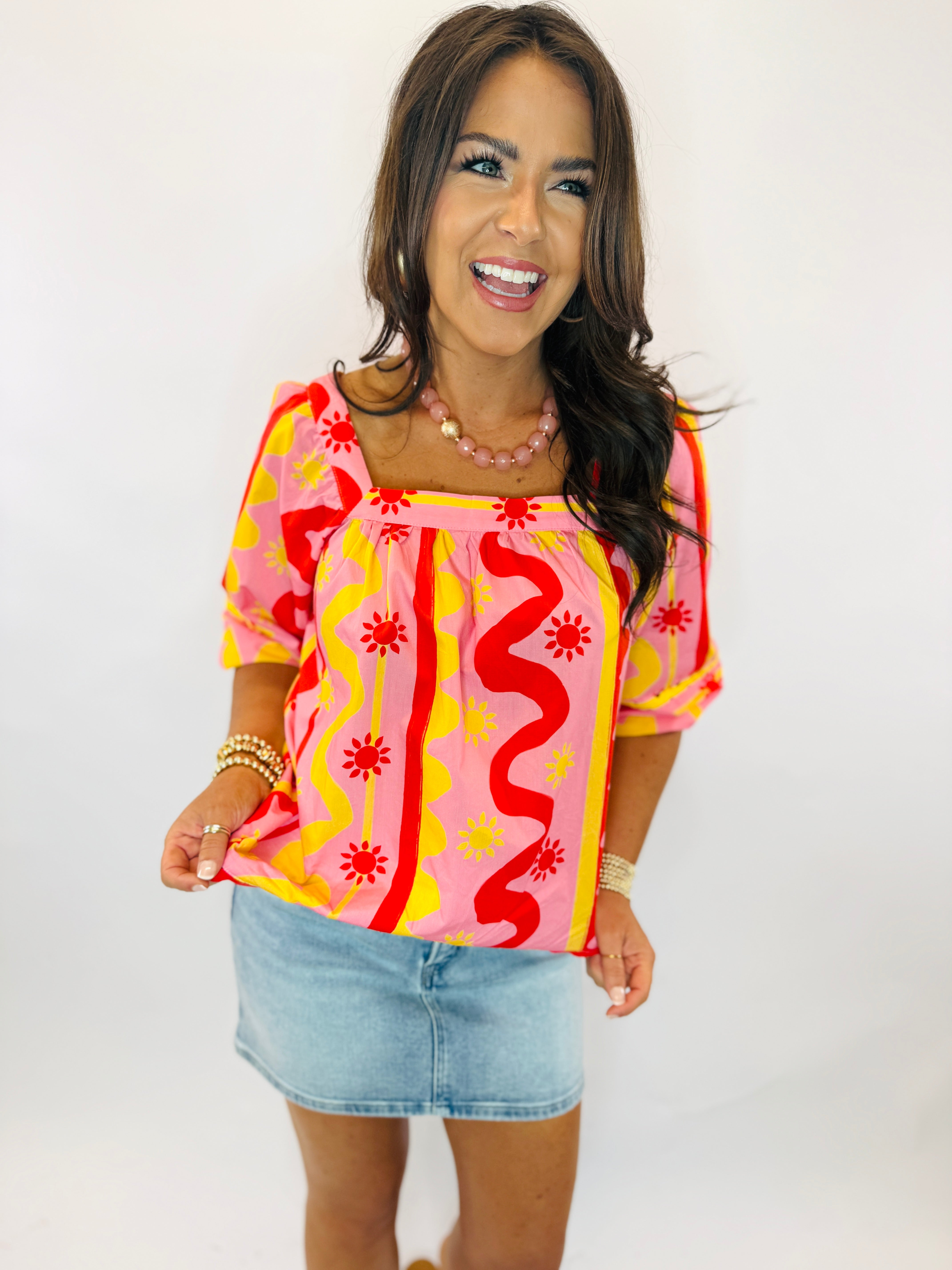 Woman wearing colorful Emmie Top with pink, red, and yellow wavy pattern paired with denim mini skirt