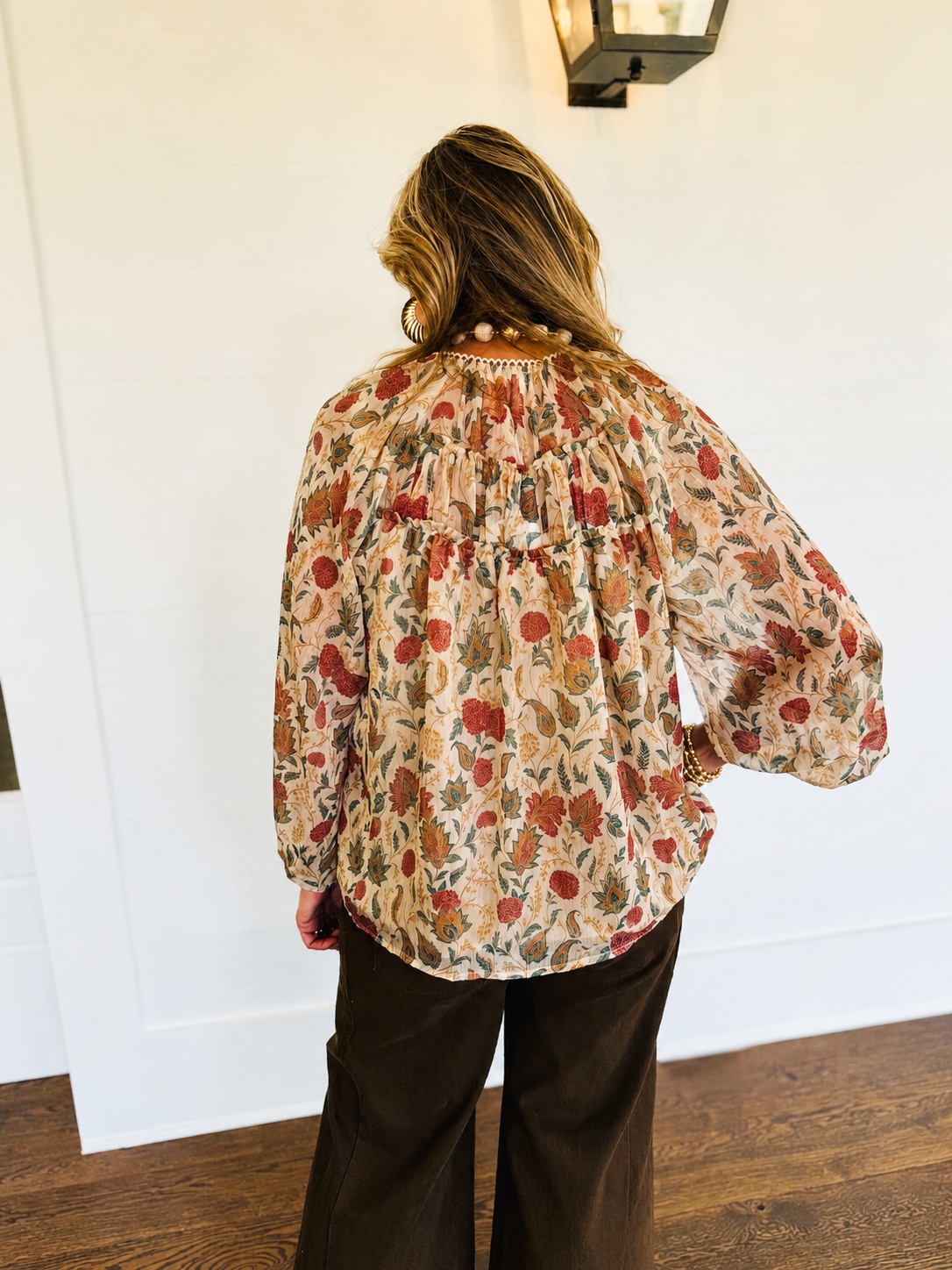 Back view of the Ella ecru multicolor floral paisley blouse, showing gathered detailing and a curved hem, styled with brown pants.