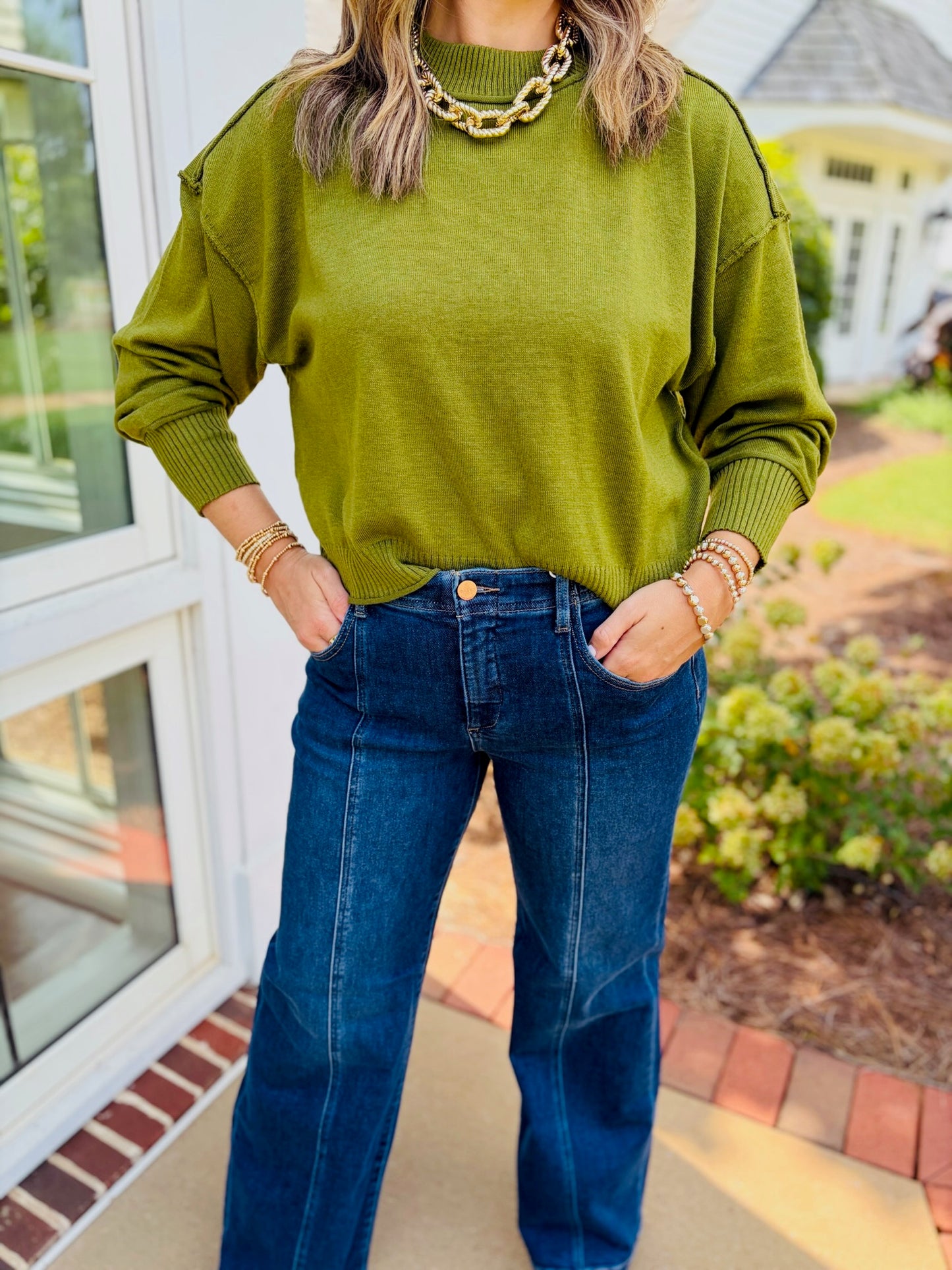 Close-up of the Emma olive crew neck sweater, showing dropped shoulders and ribbed neckline, cuffs, and hem, styled with blue jeans.