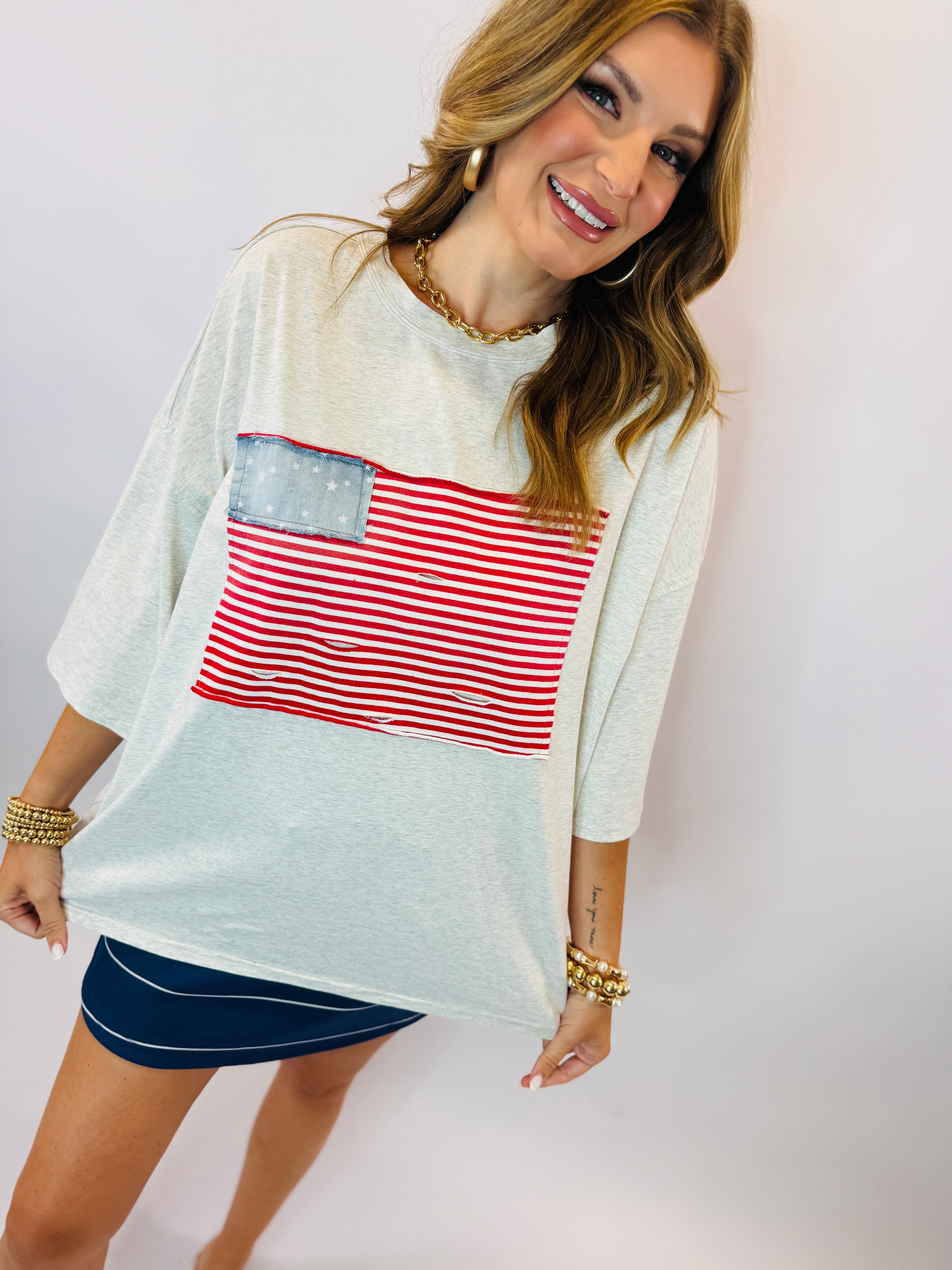Woman wearing white oversized tee with red striped American flag design and navy shorts, smiling at camera.