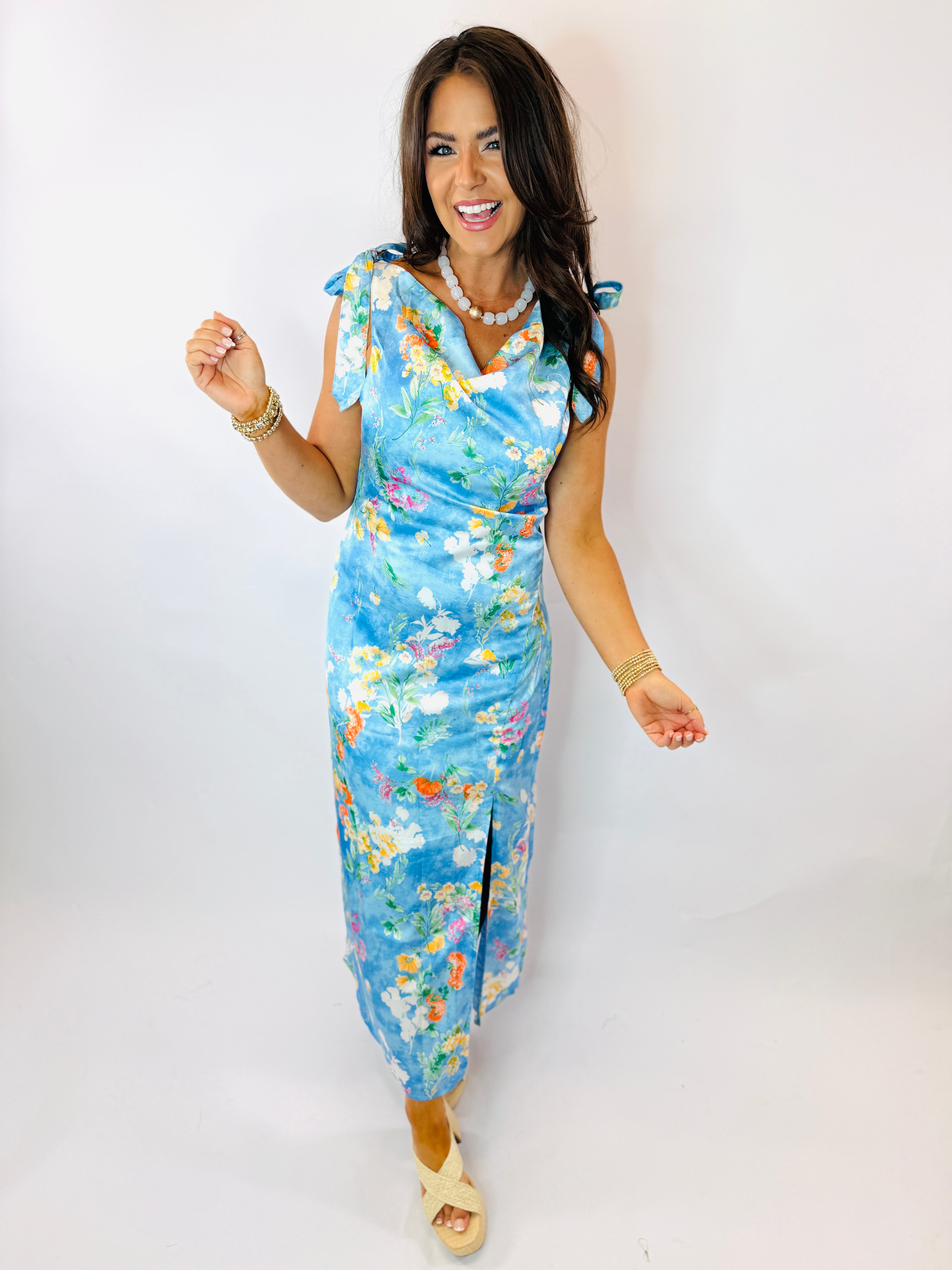 Woman wearing blue floral wrap dress with tie shoulders, pearl necklace, and gold accessories against white background.