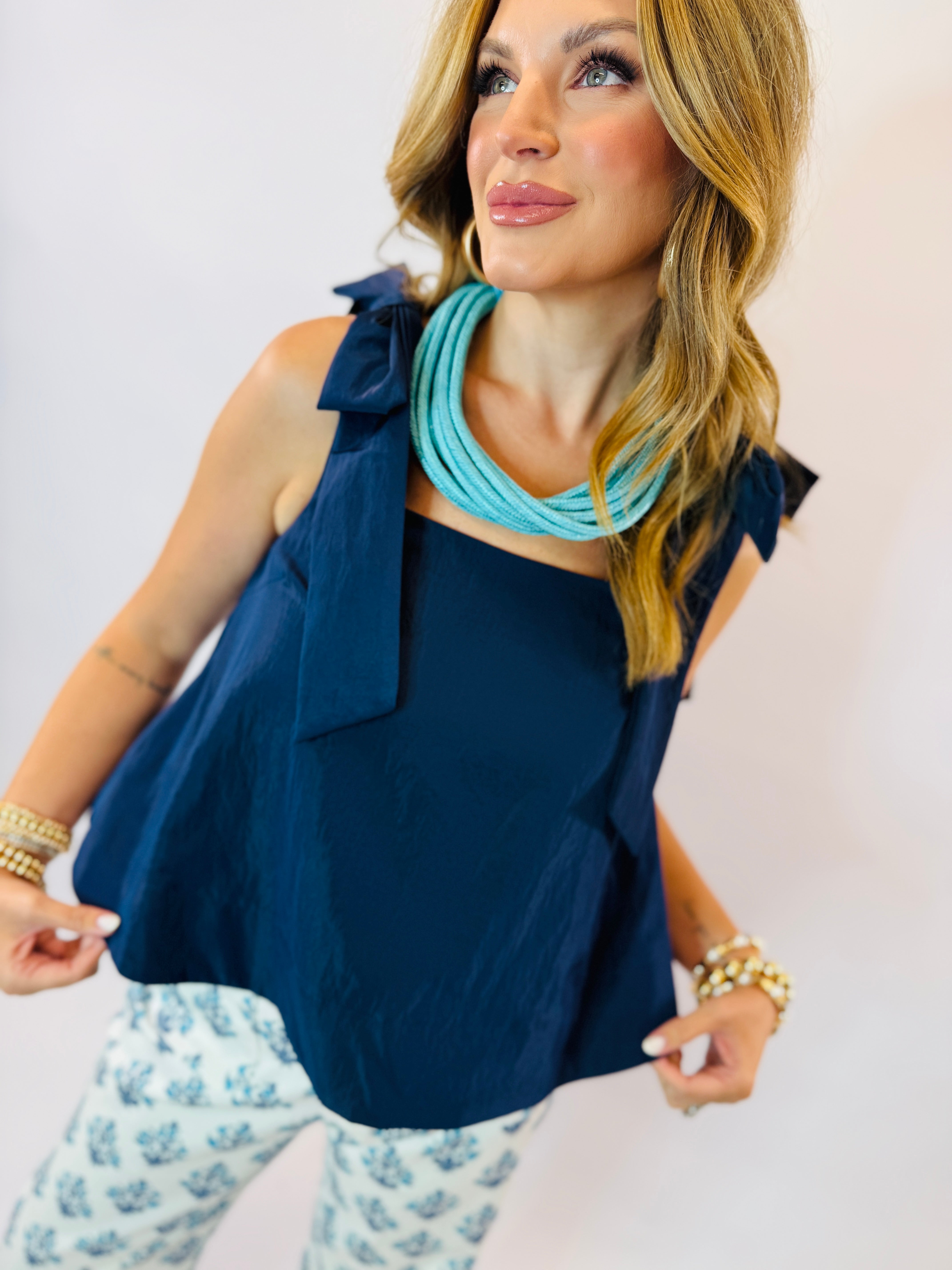 Woman wearing navy blue sleeveless tank top with bow detail on shoulder, paired with turquoise necklace and patterned bott...