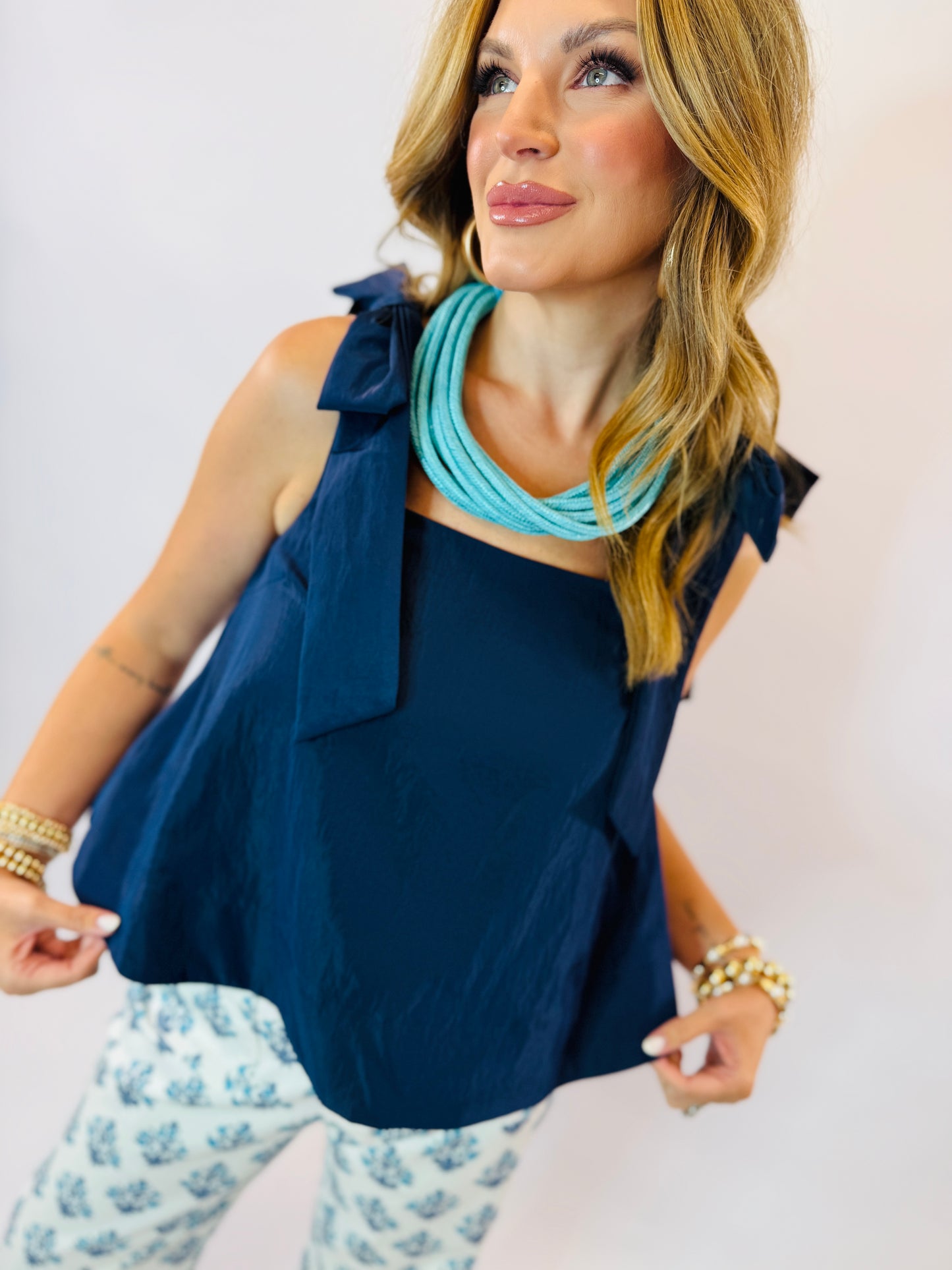 Woman wearing navy blue sleeveless tank top with bow detail on shoulder, paired with turquoise necklace and patterned bott...