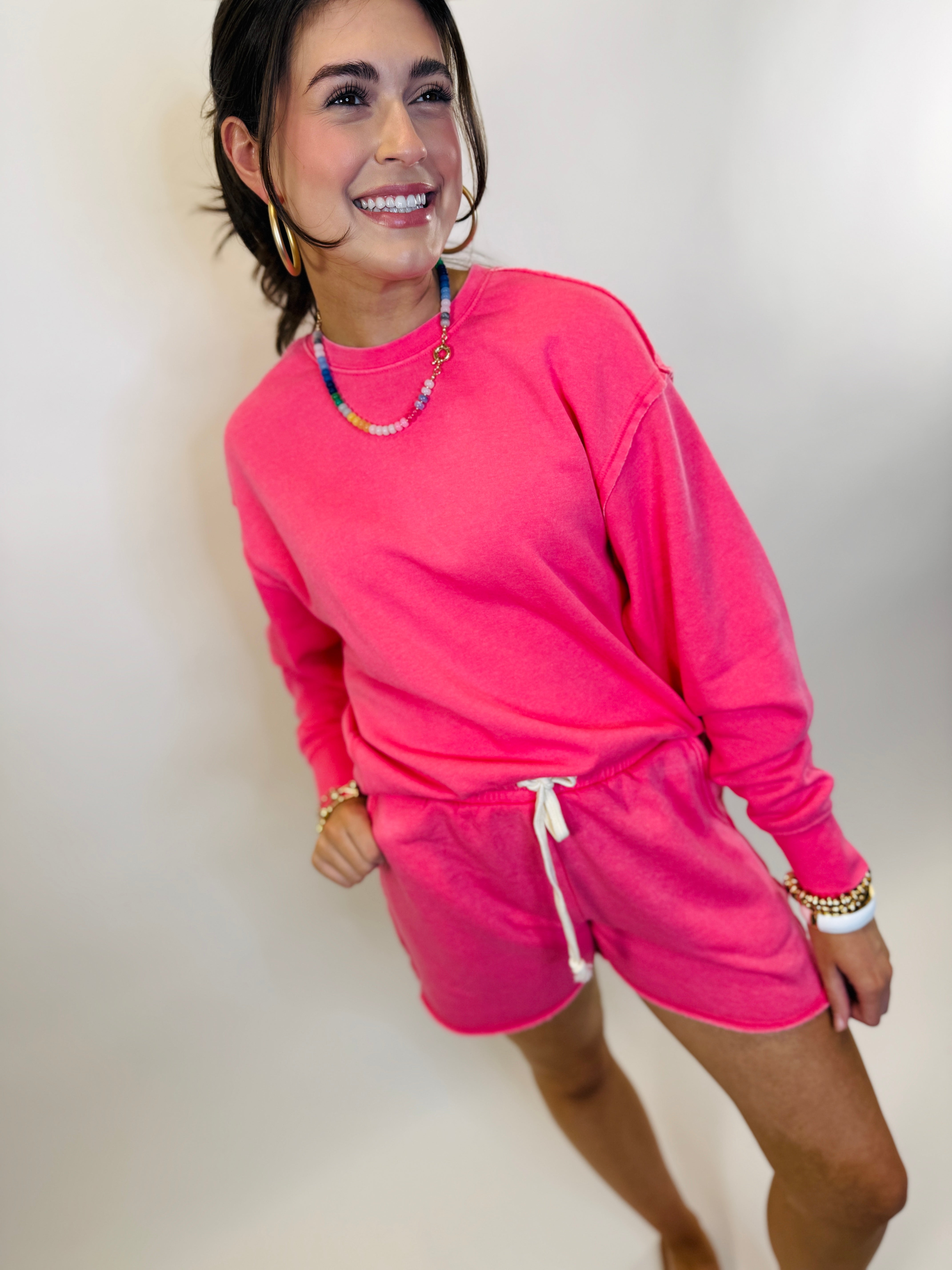 Woman wearing bright pink sweatshirt and matching shorts set with colorful beaded necklace and gold jewelry accessories.