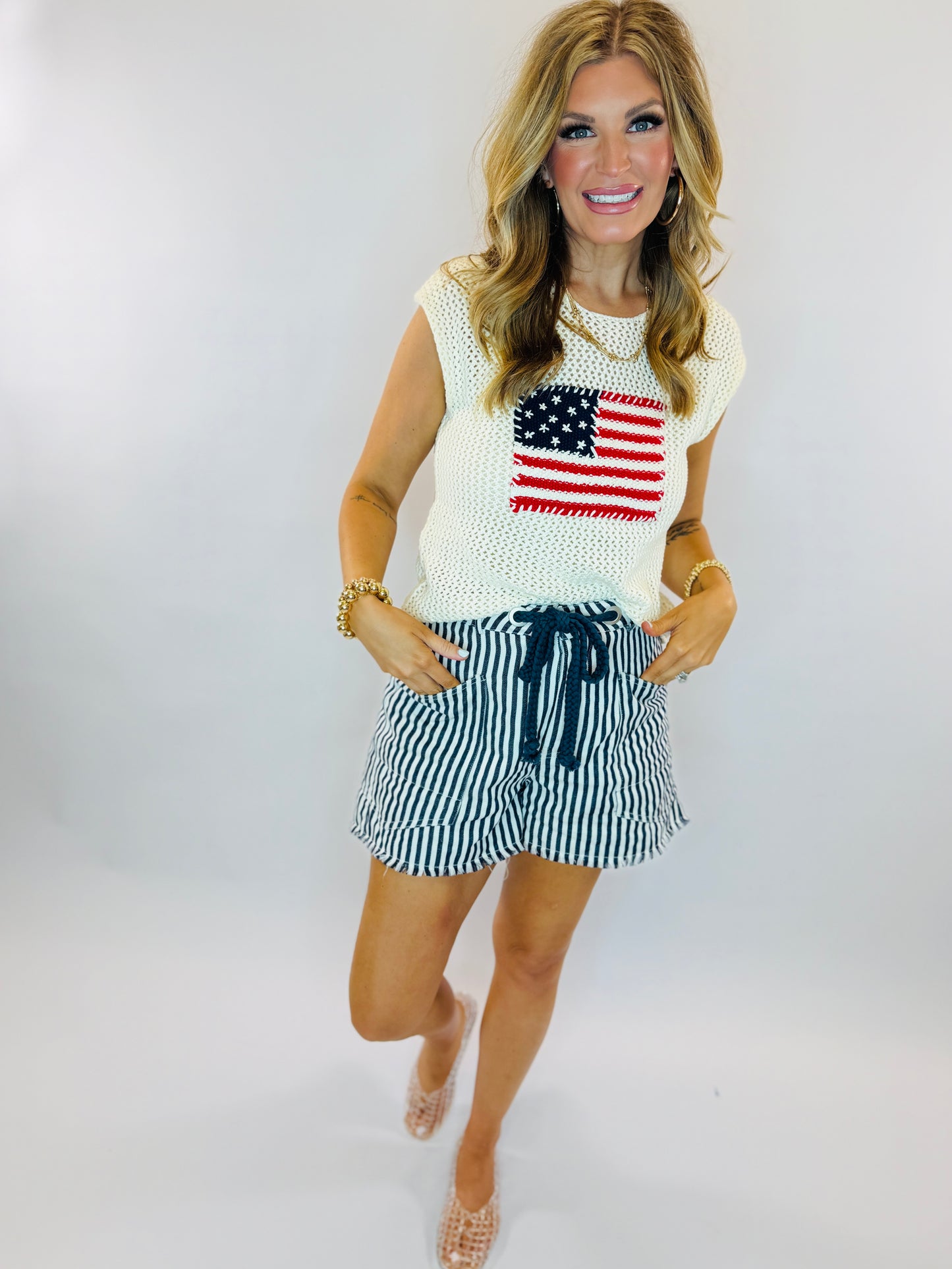 Woman wearing white sleeveless American flag sweater and navy striped drawstring shorts with beaded sandals