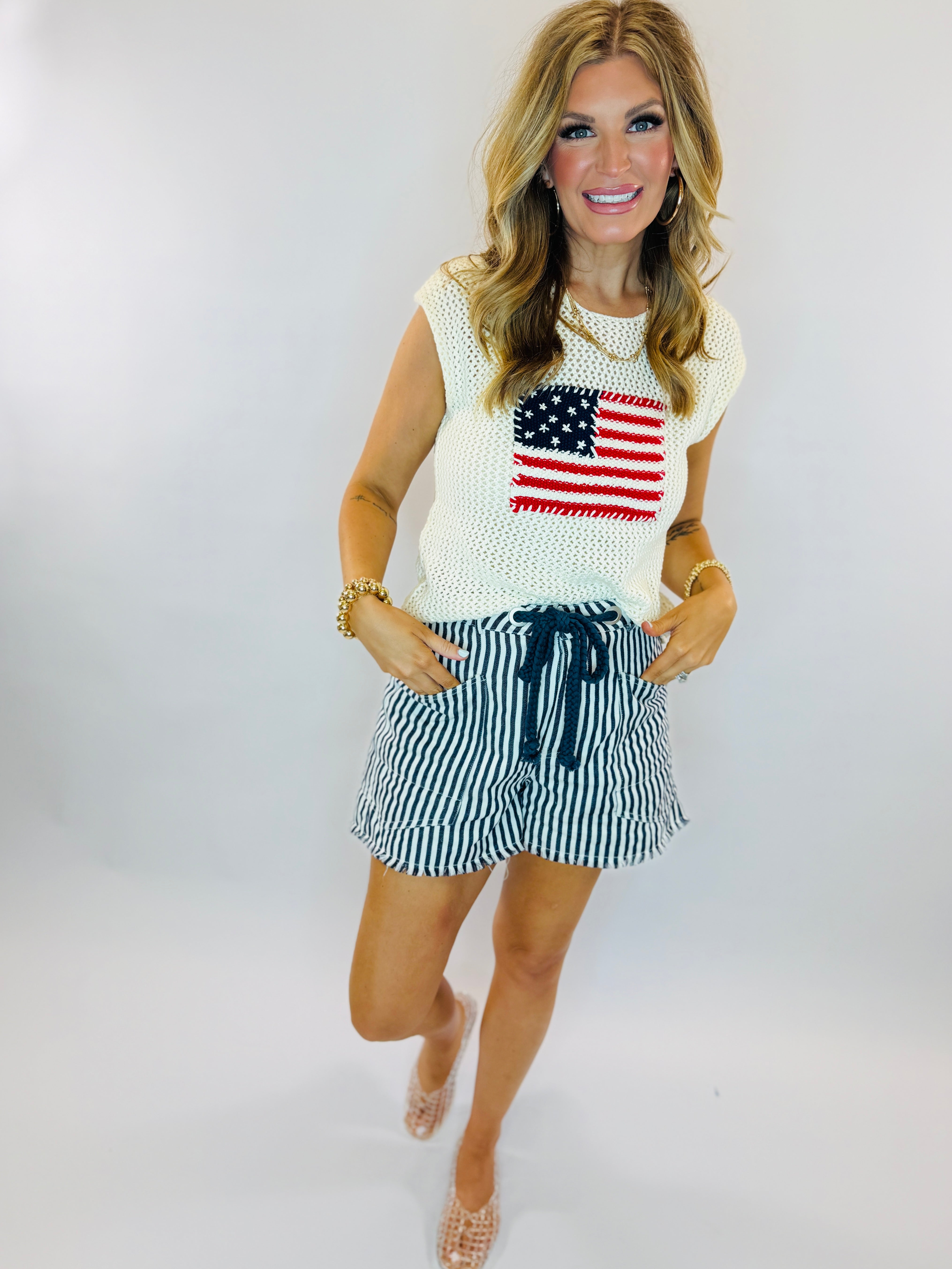Woman wearing white sleeveless American flag sweater and navy striped drawstring shorts with beaded sandals
