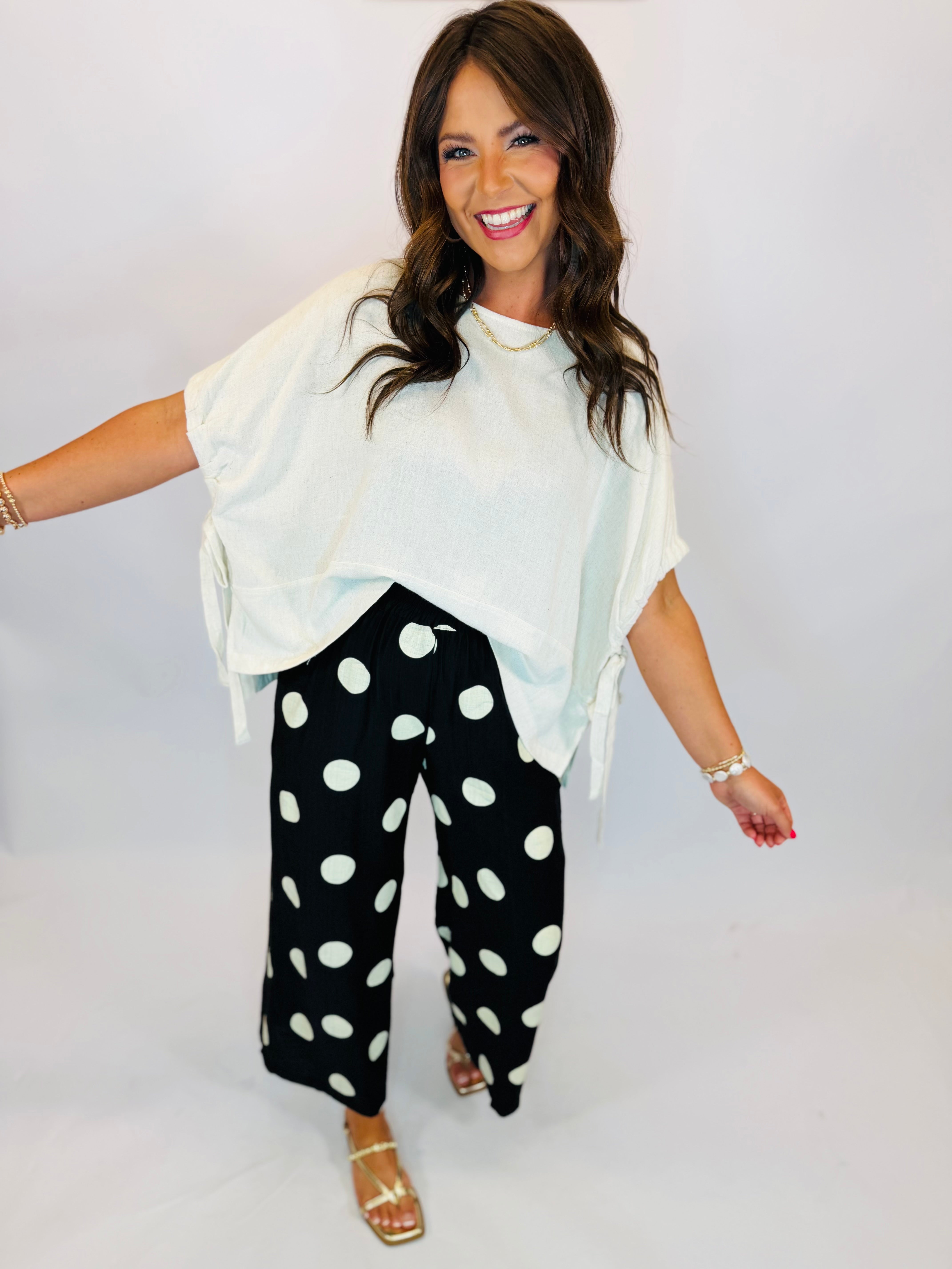 Woman wearing a white boxy short-sleeve Cici Top with side tie details, paired with black polka dot pants.