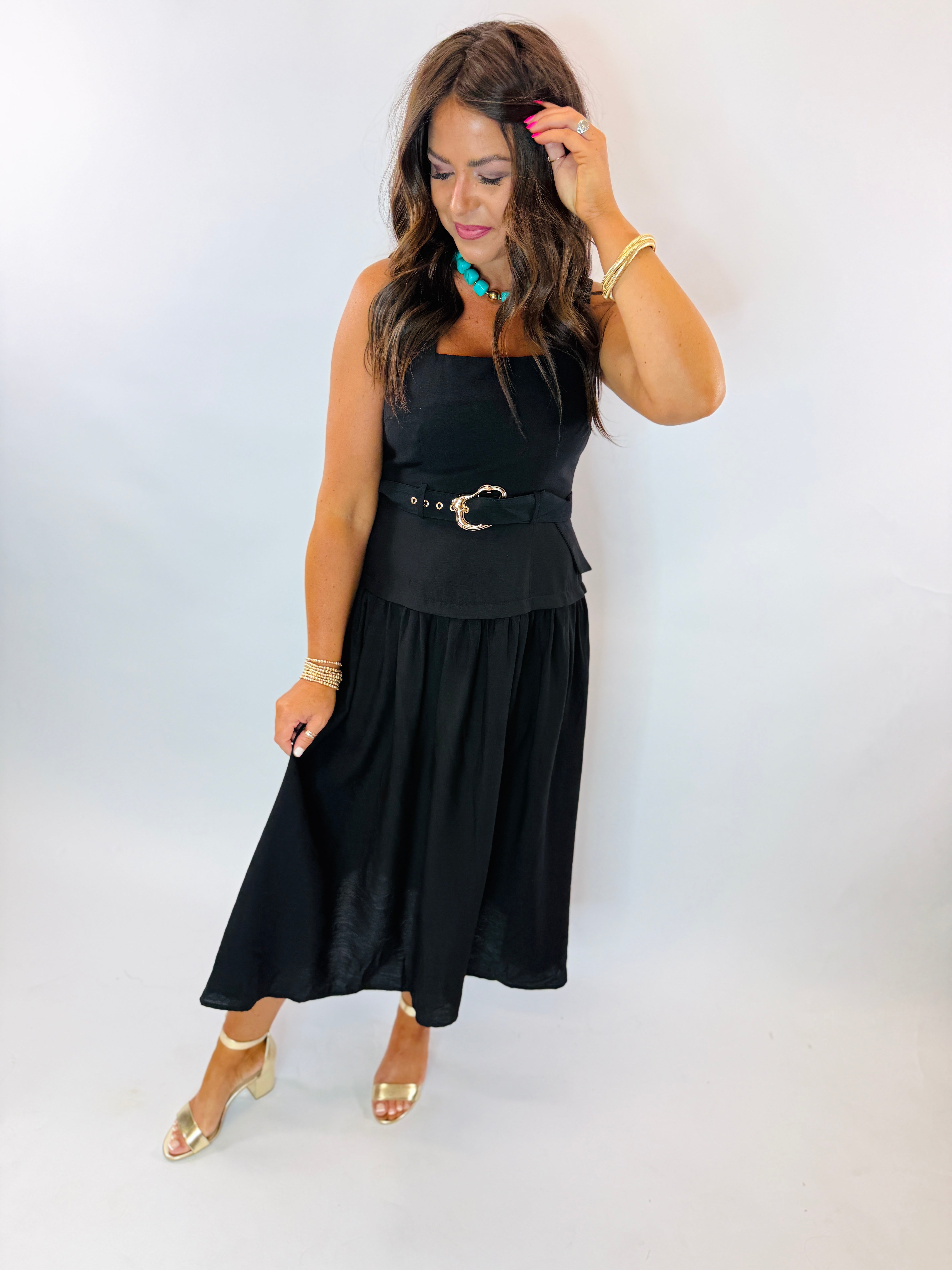 Woman wearing black sleeveless midi dress with gold belt buckle, turquoise necklace, and gold heels.