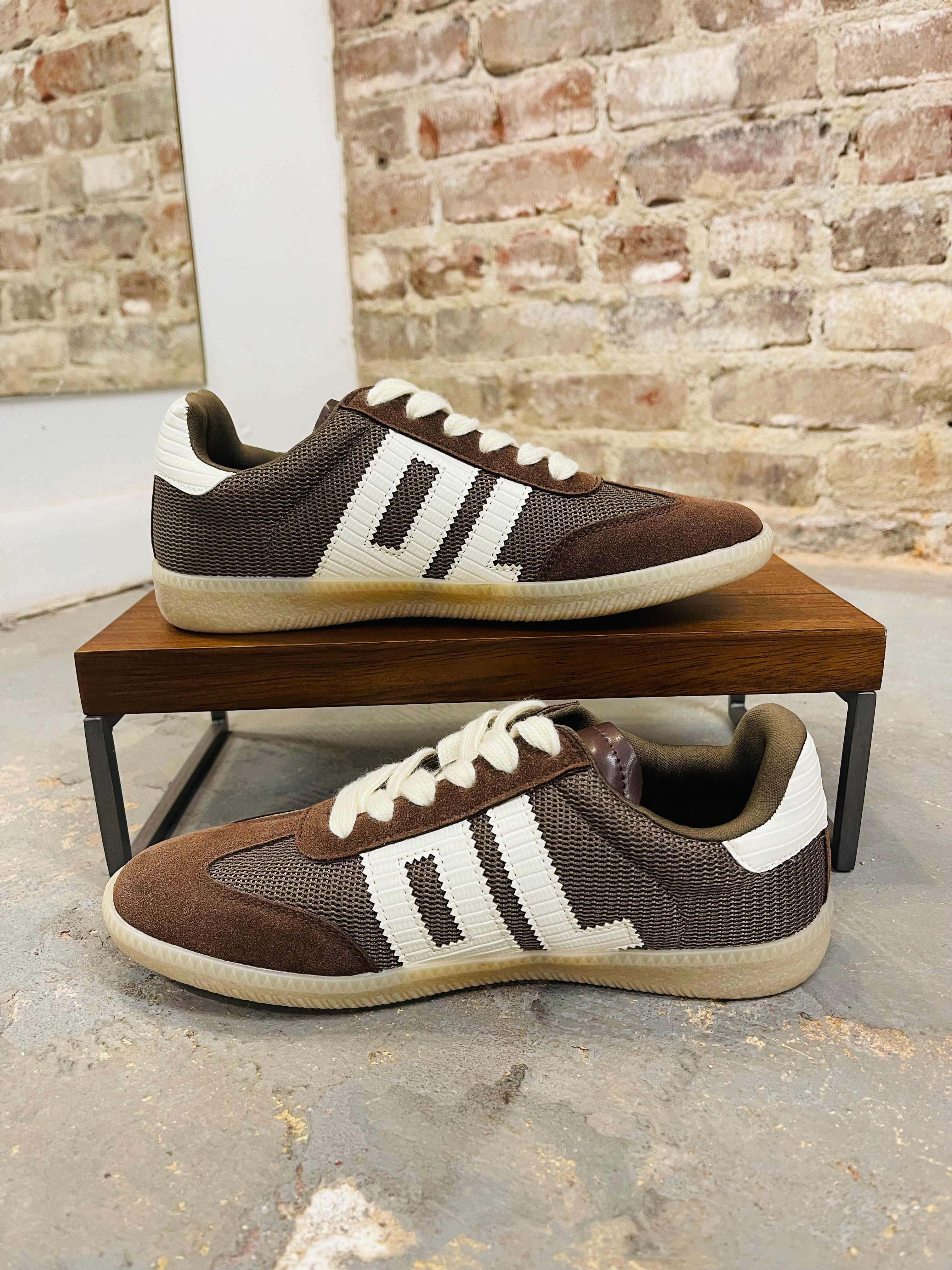 Brown Emma sneakers with white three-stripe design and cream soles displayed on wooden bench against brick wall