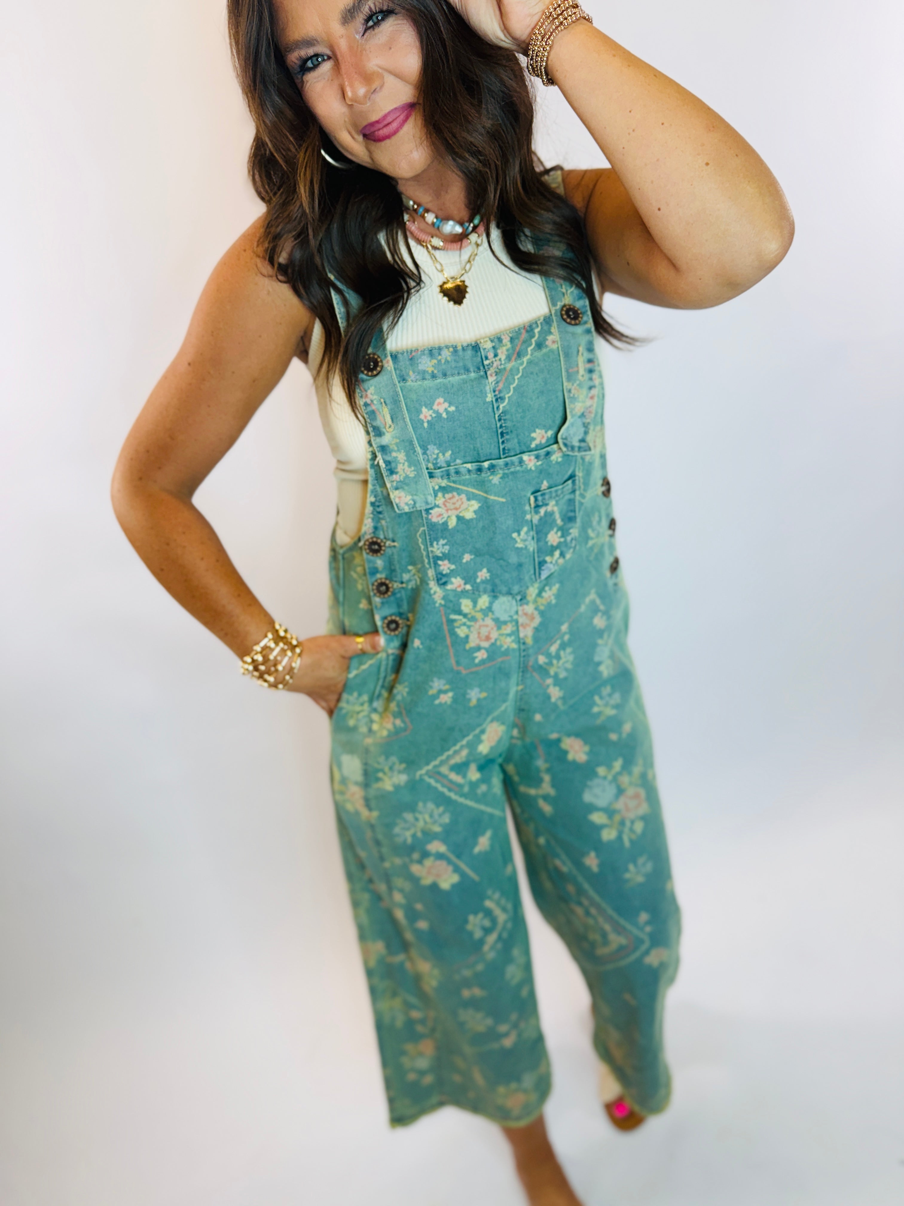 Woman wearing teal floral print denim cropped overalls with white bodysuit and gold accessories.