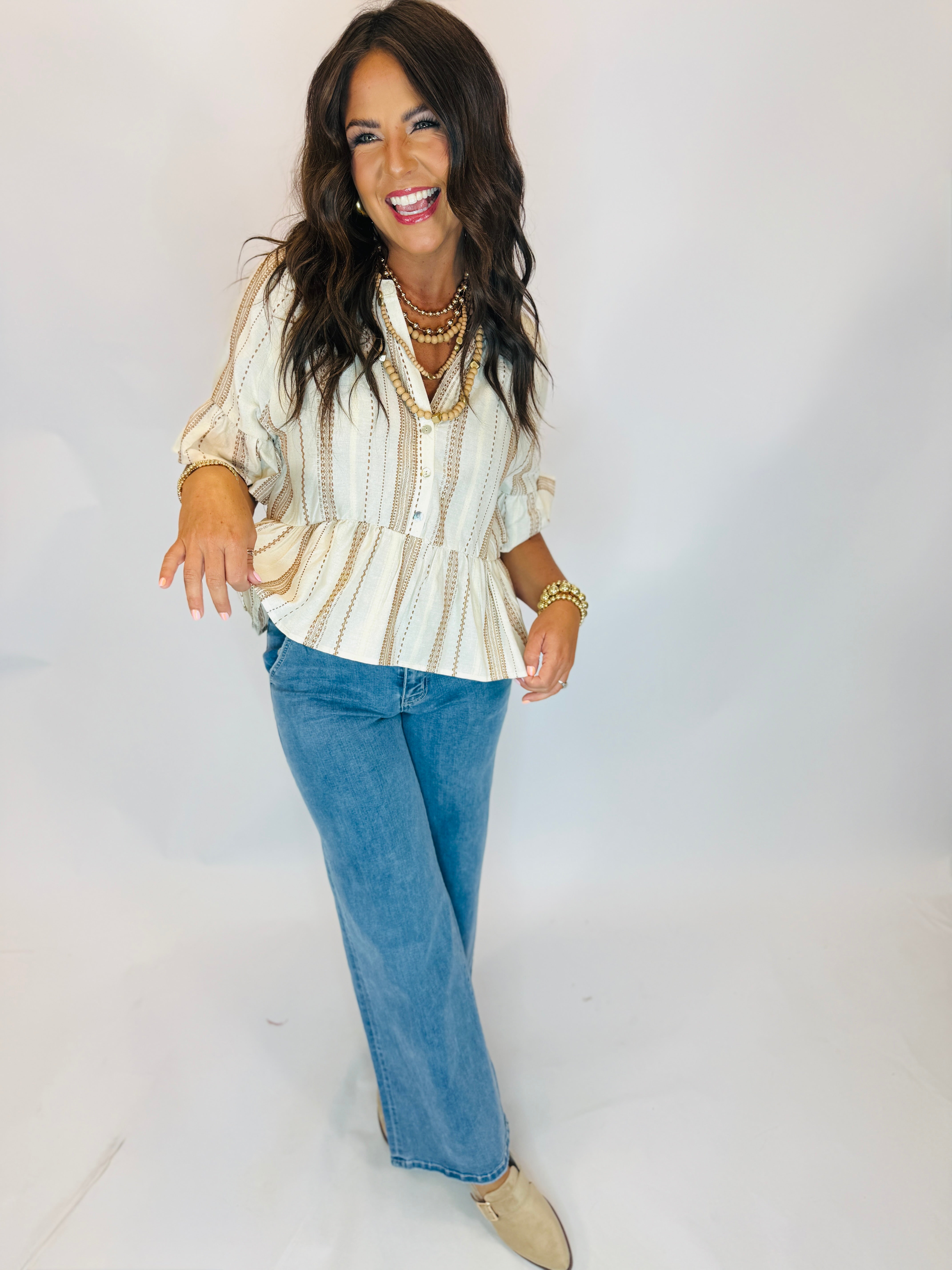 Woman wearing Charlie Too cream striped peplum blouse with brown embroidery, paired with wide-leg jeans and layered beaded...