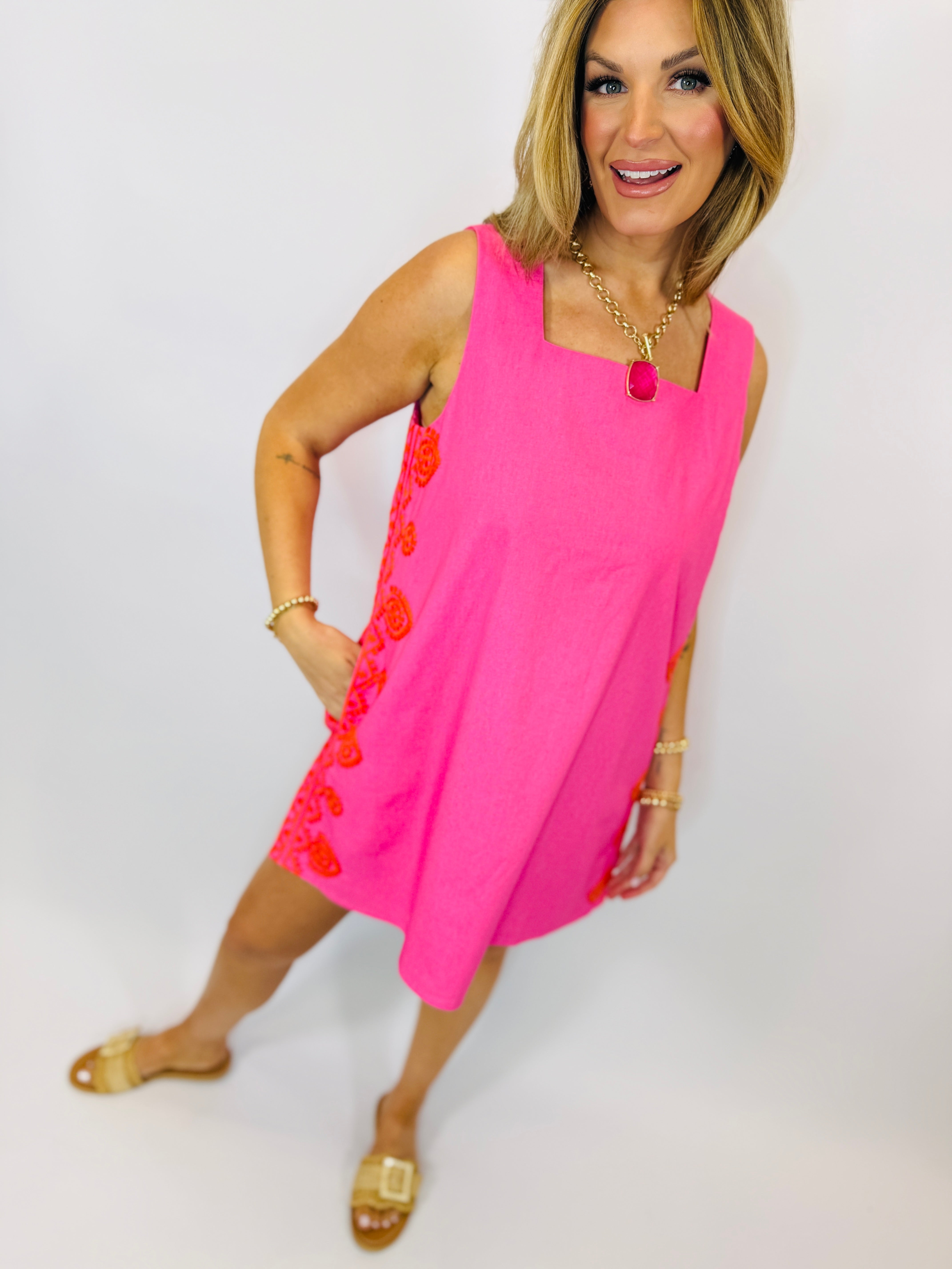 Woman wearing bright pink sleeveless Marlene dress with red floral embroidery detail and gold sandals