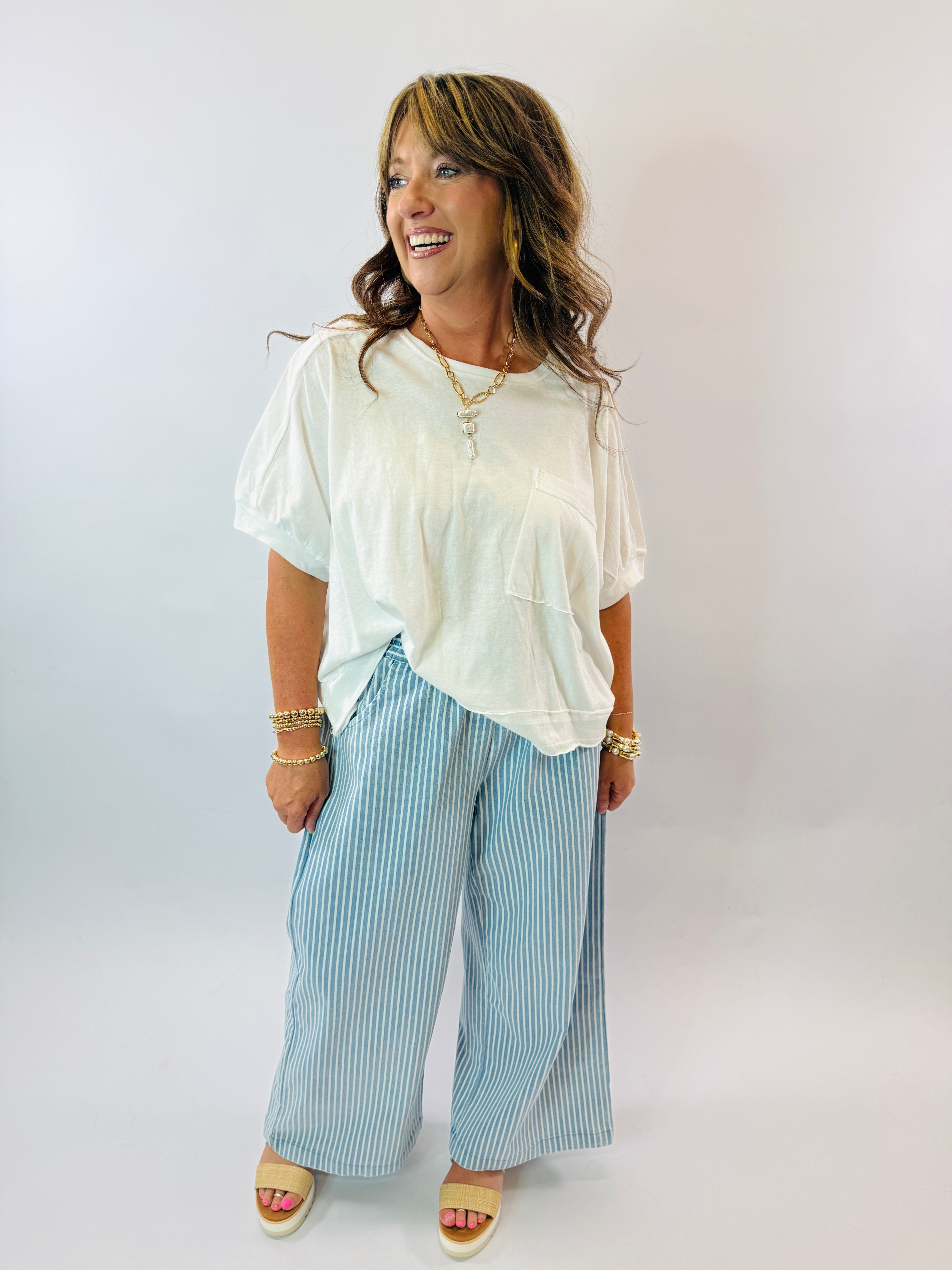 Woman wearing white oversized top and blue striped wide-leg pants with gold accessories and sandals.