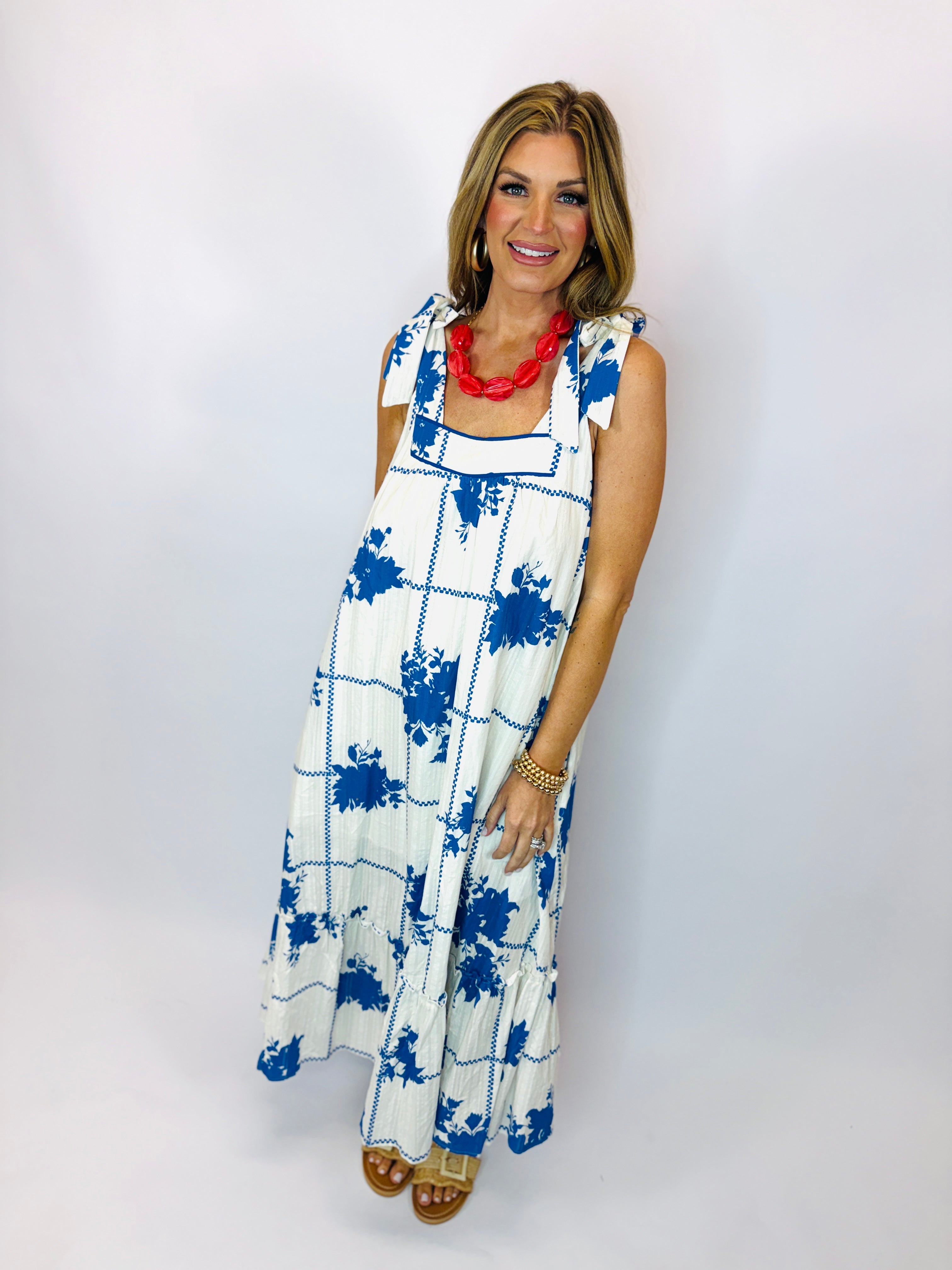 Woman wearing white maxi dress with blue floral print, red statement necklace, and brown sandals.