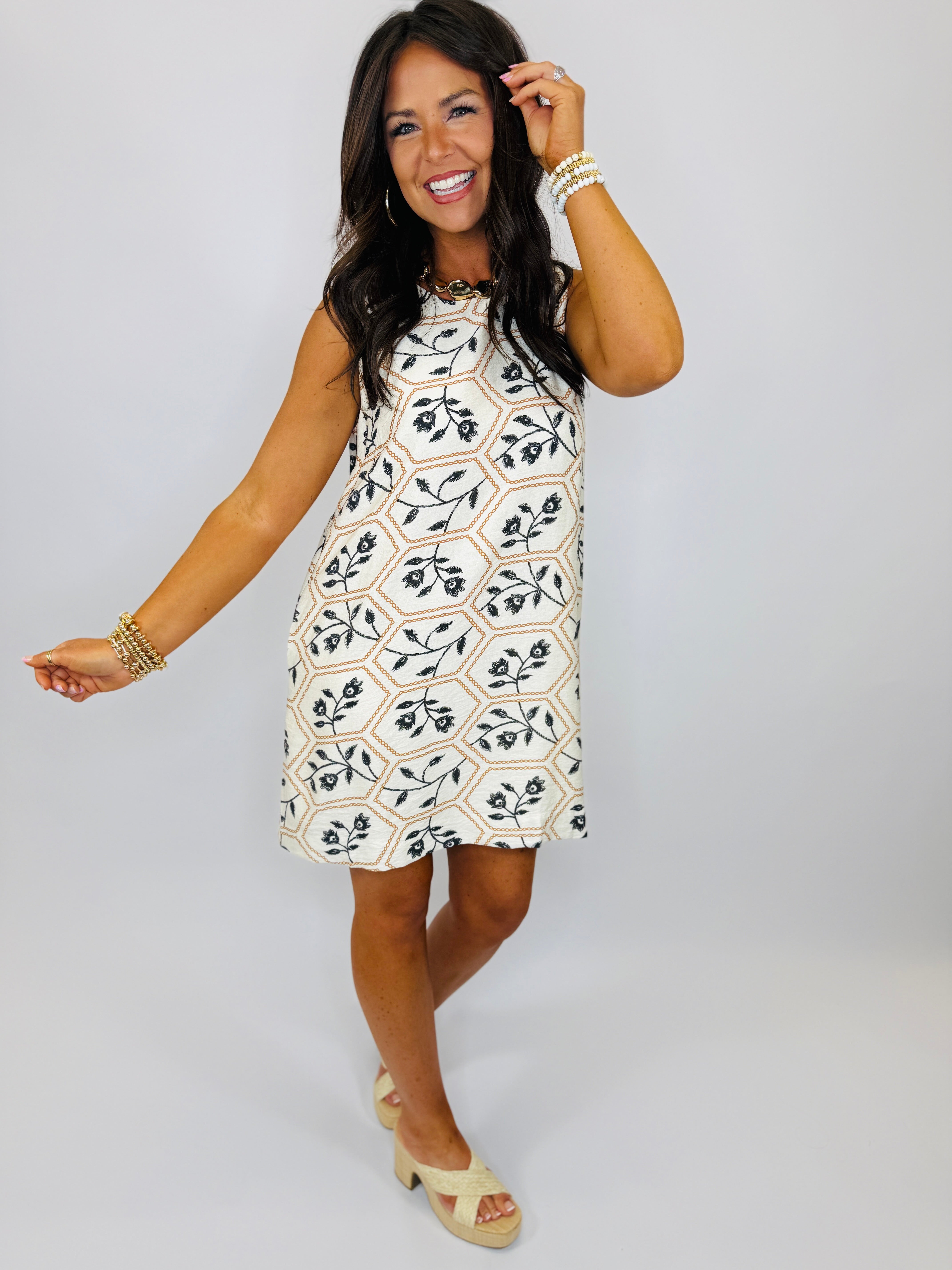 Woman wearing white sleeveless Christina dress with geometric floral pattern, styled with gold jewelry and platform sandals.