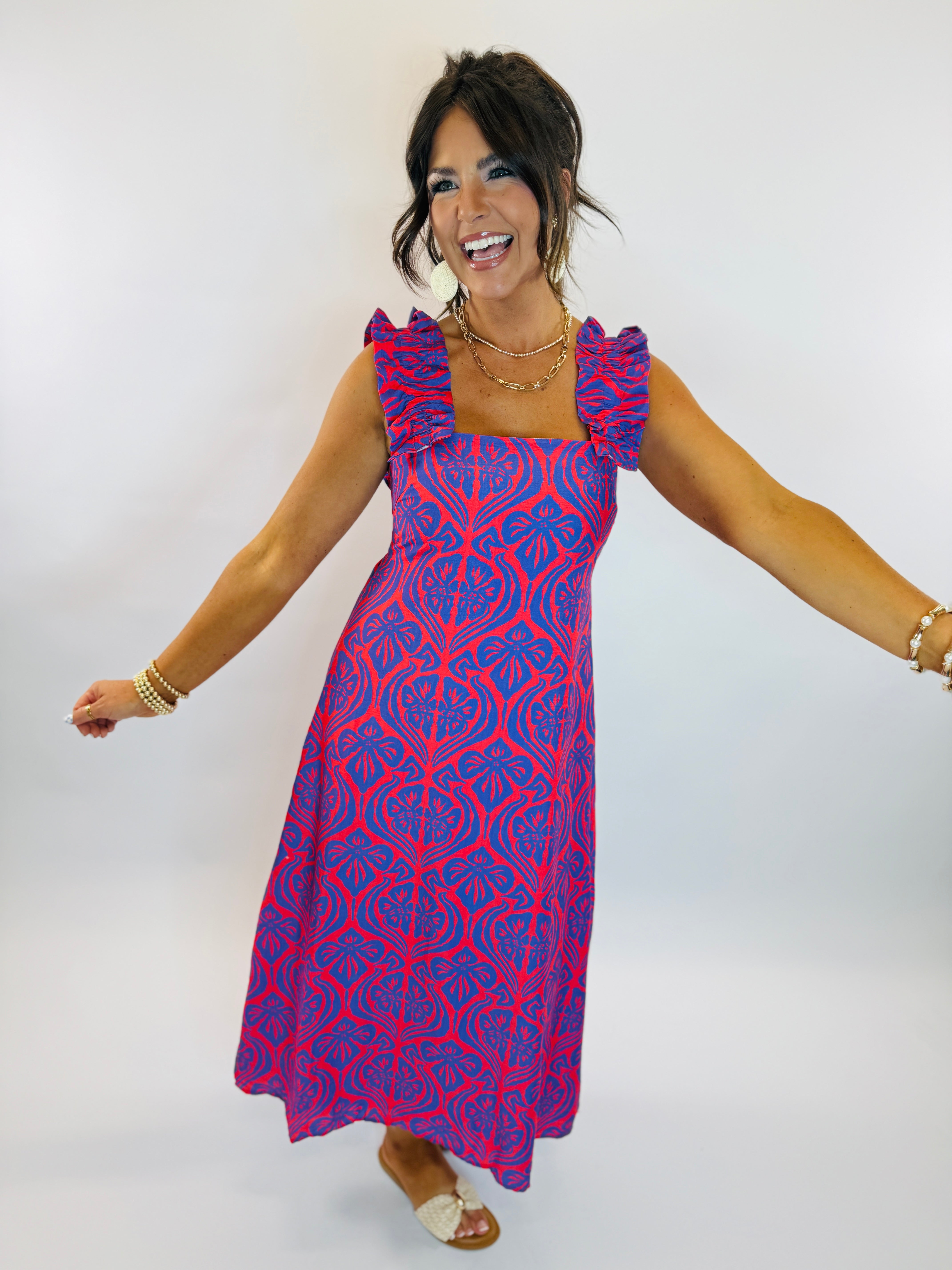 Woman wearing vibrant red and blue patterned Ciara dress with ruffle straps, smiling with arms outstretched.