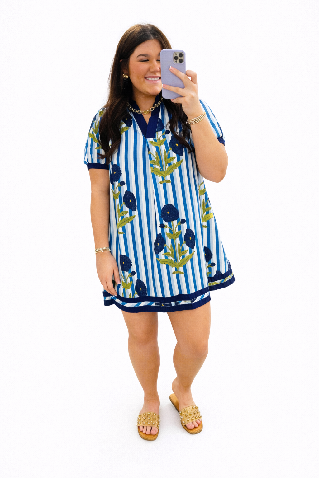 Woman wearing blue and white striped dress with floral print, taking mirror selfie with studded gold sandals.