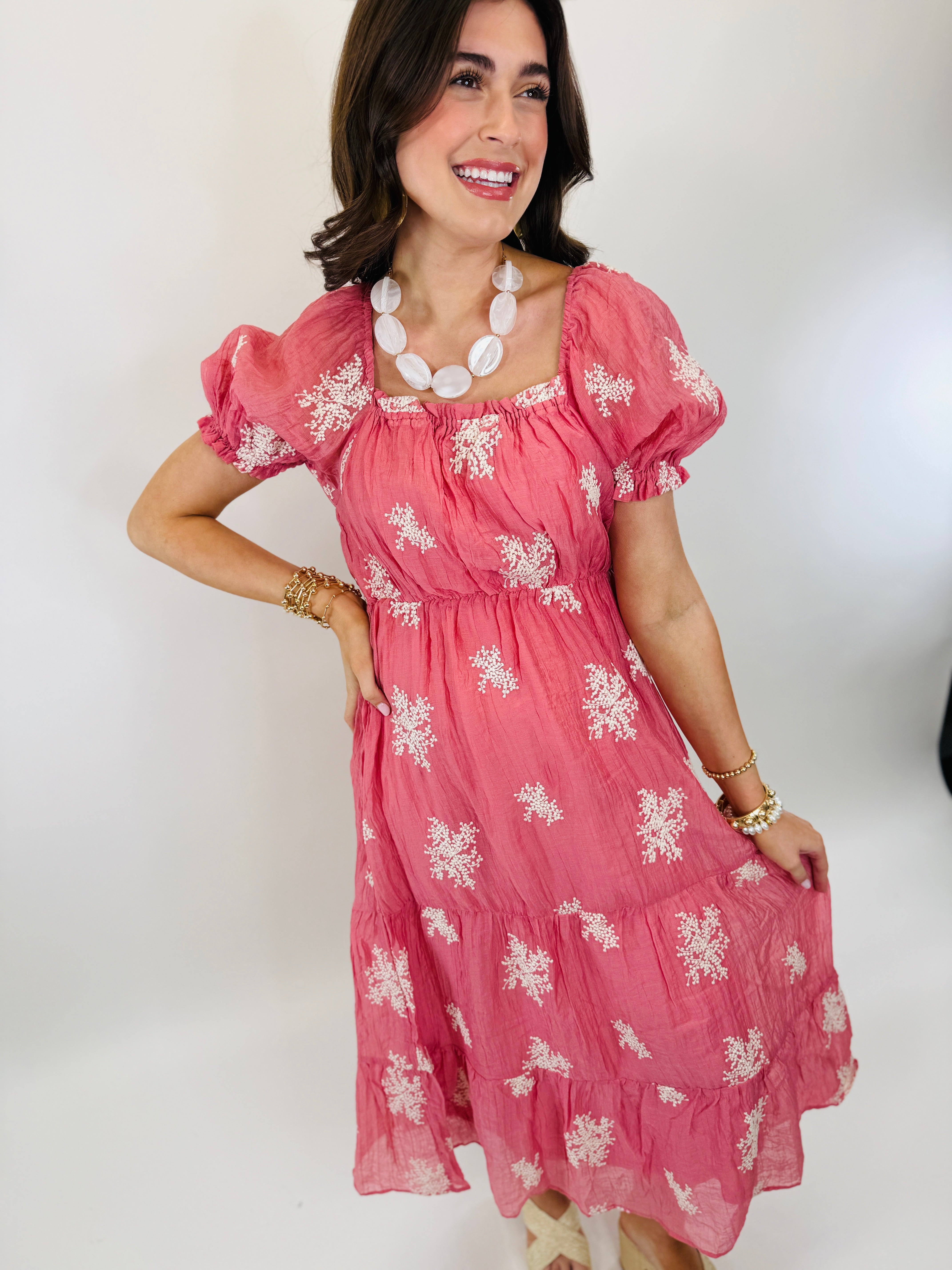 Pink floral midi dress with puffed sleeves and tiered ruffles, styled with white beaded necklace and gold bracelets.
