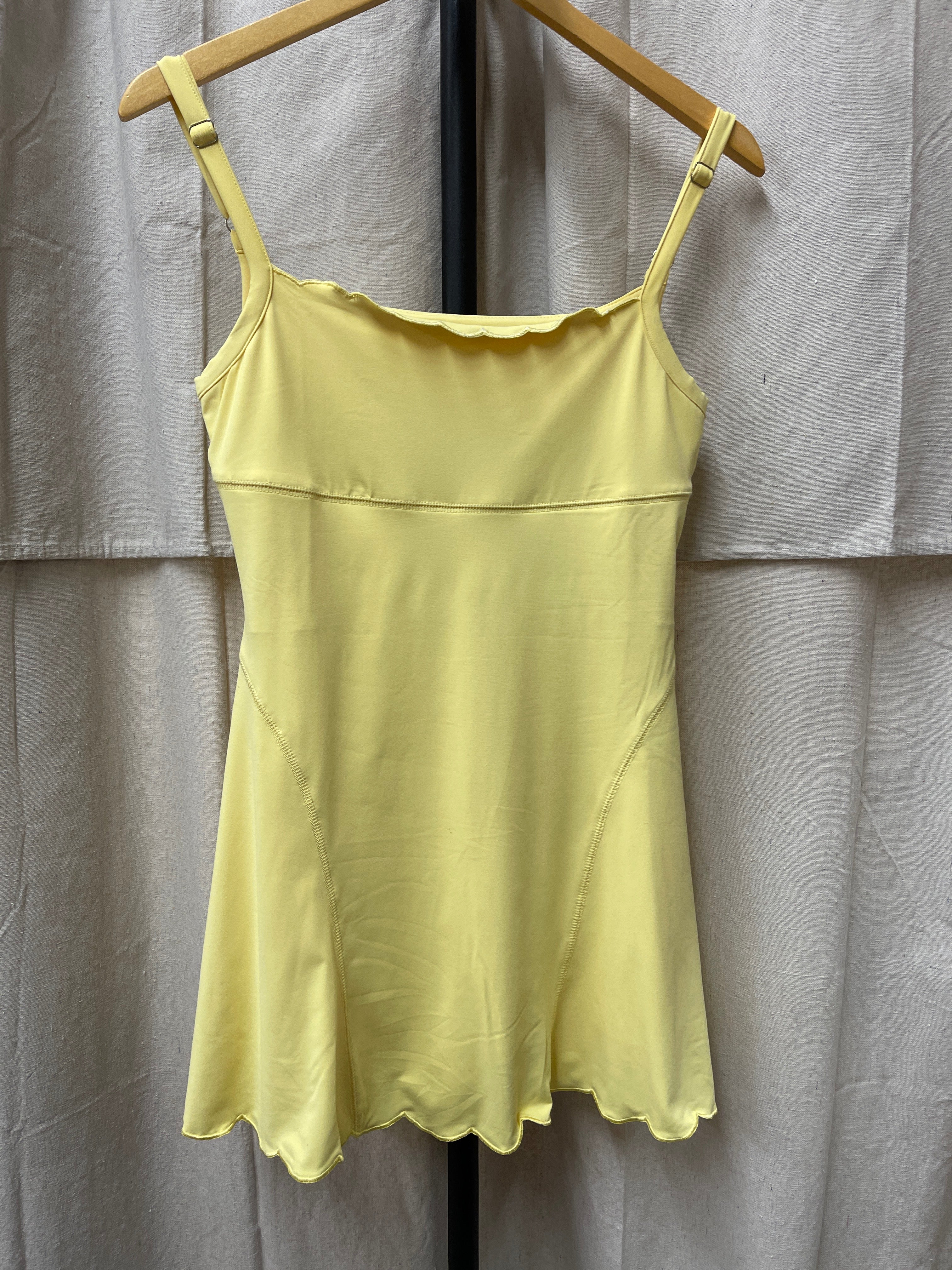 Yellow sleeveless mini dress with adjustable straps and scalloped hem hanging on wooden hanger