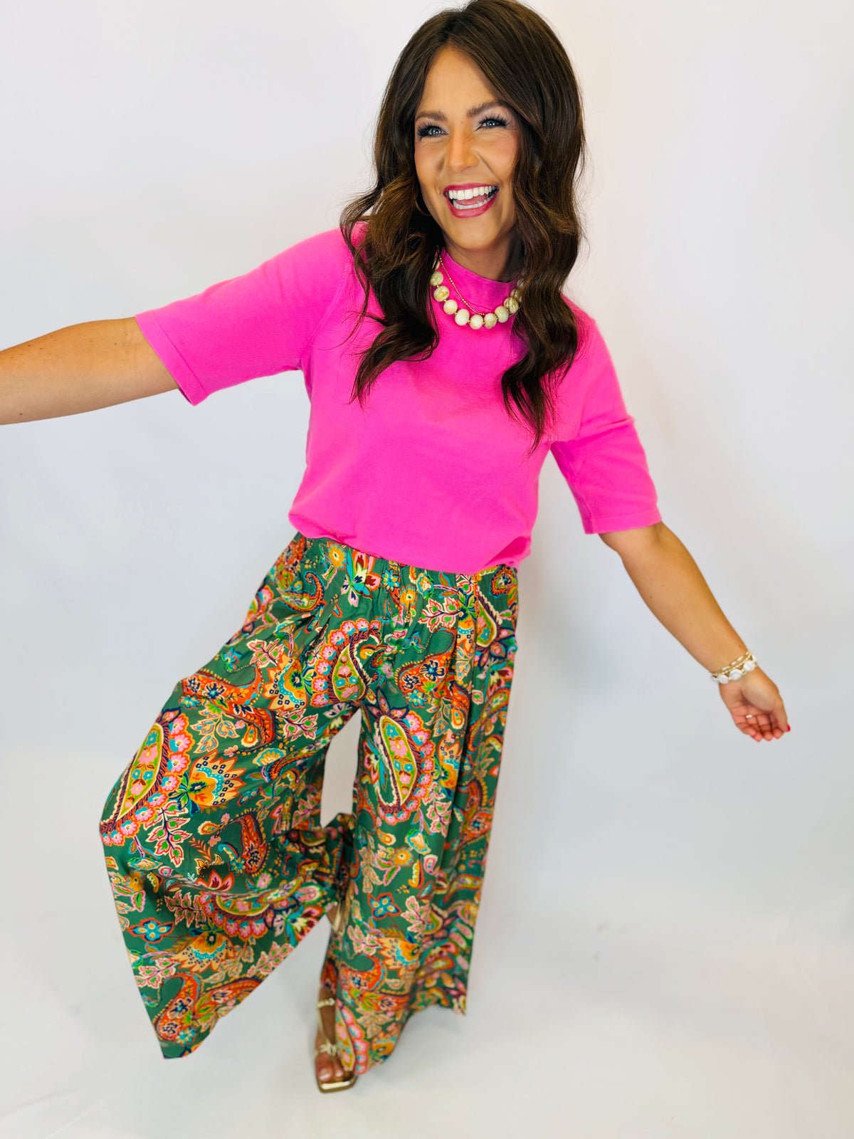 Woman wearing bright pink short-sleeve Angela Sweater Top paired with green paisley wide-leg pants and pearl necklace.