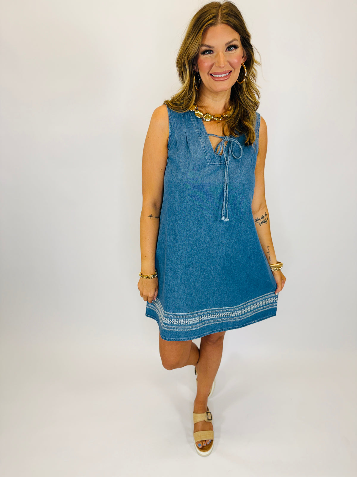 Woman wearing blue denim sleeveless dress with lace-up neckline, embroidered hem detail, and platform sandals.