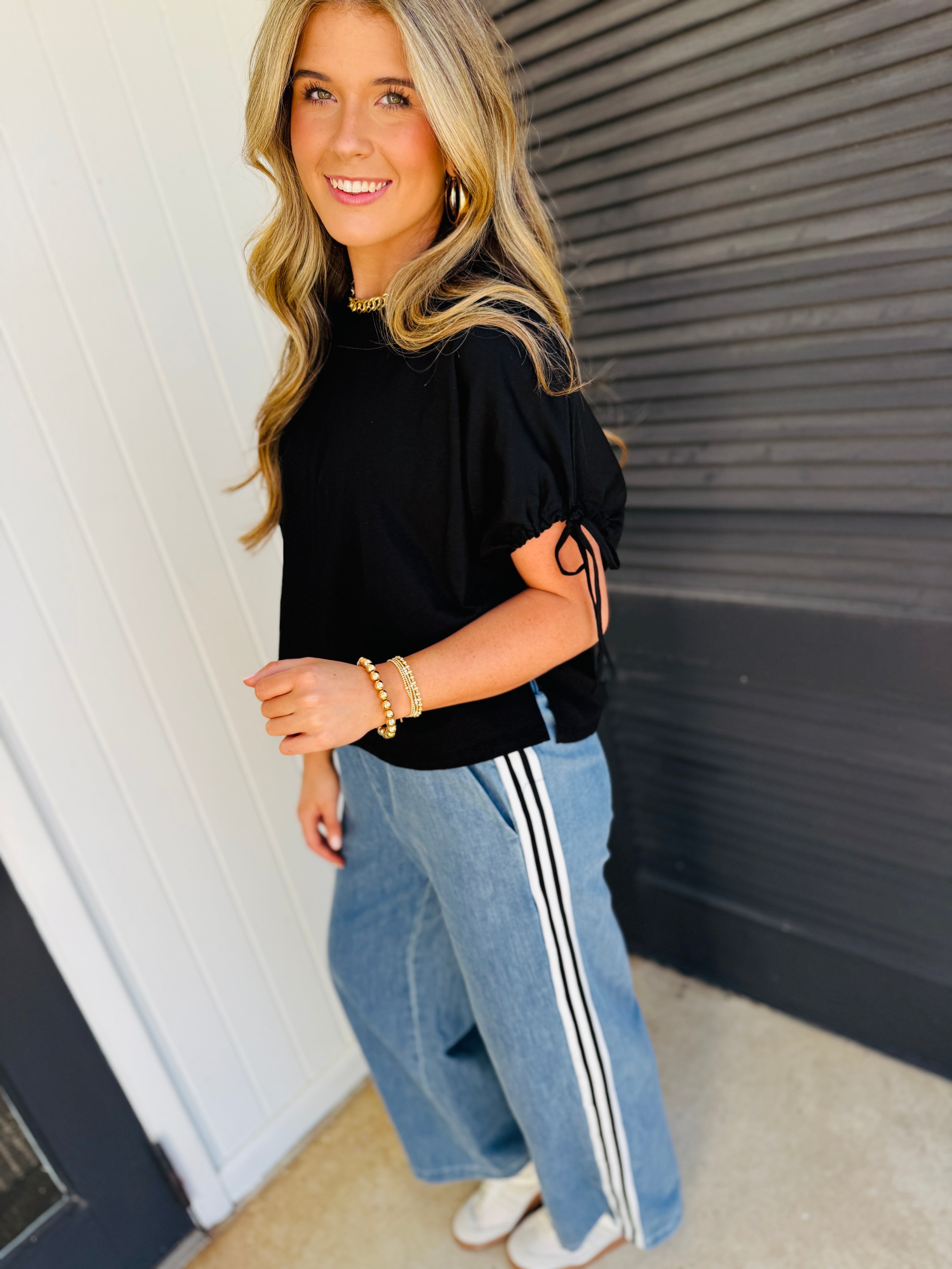 Woman wearing light blue relaxed-fit pants with white side stripes and black top, smiling at camera.