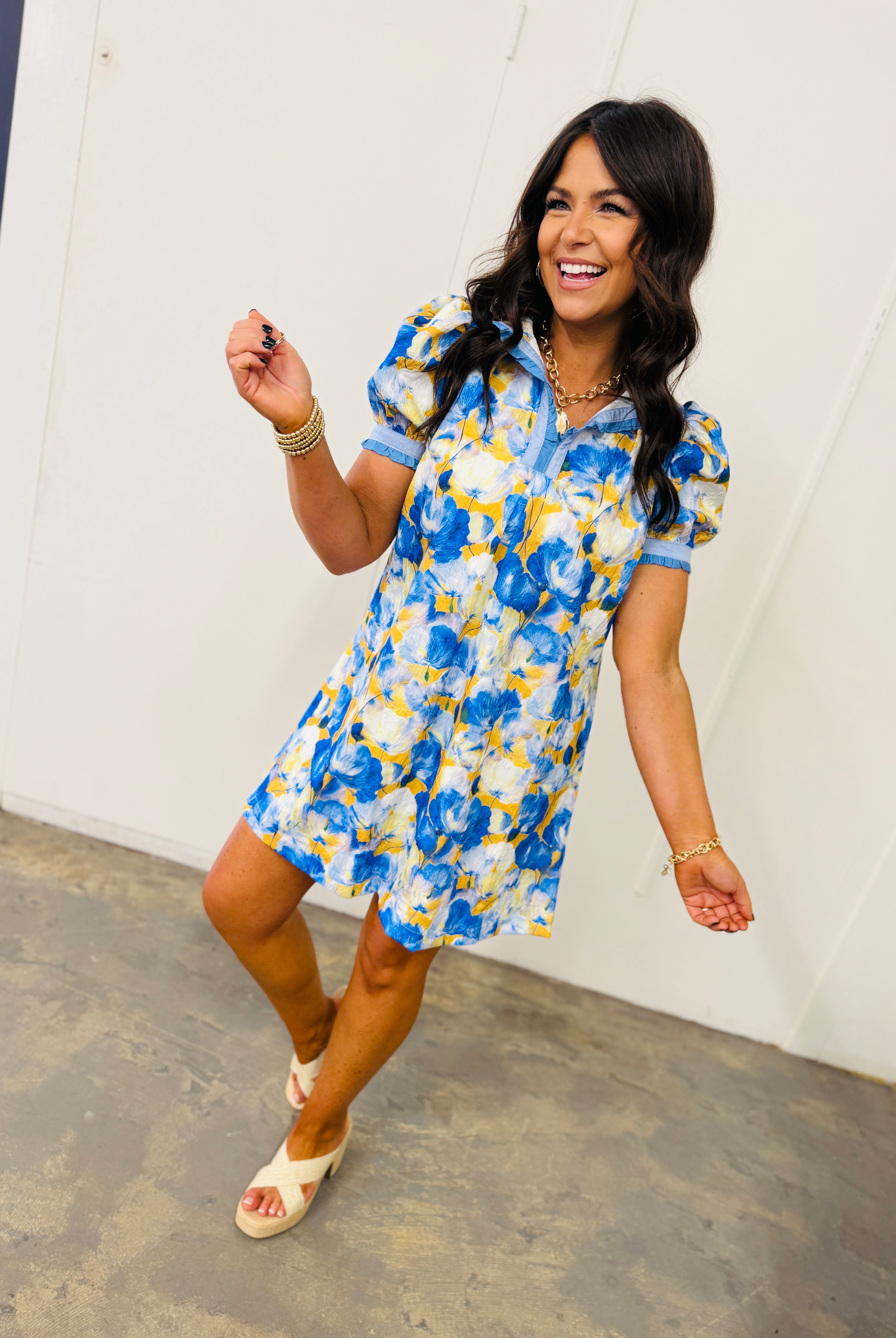 Woman wearing blue and yellow floral print short-sleeve dress with collar, styled with gold jewelry and white sandals.