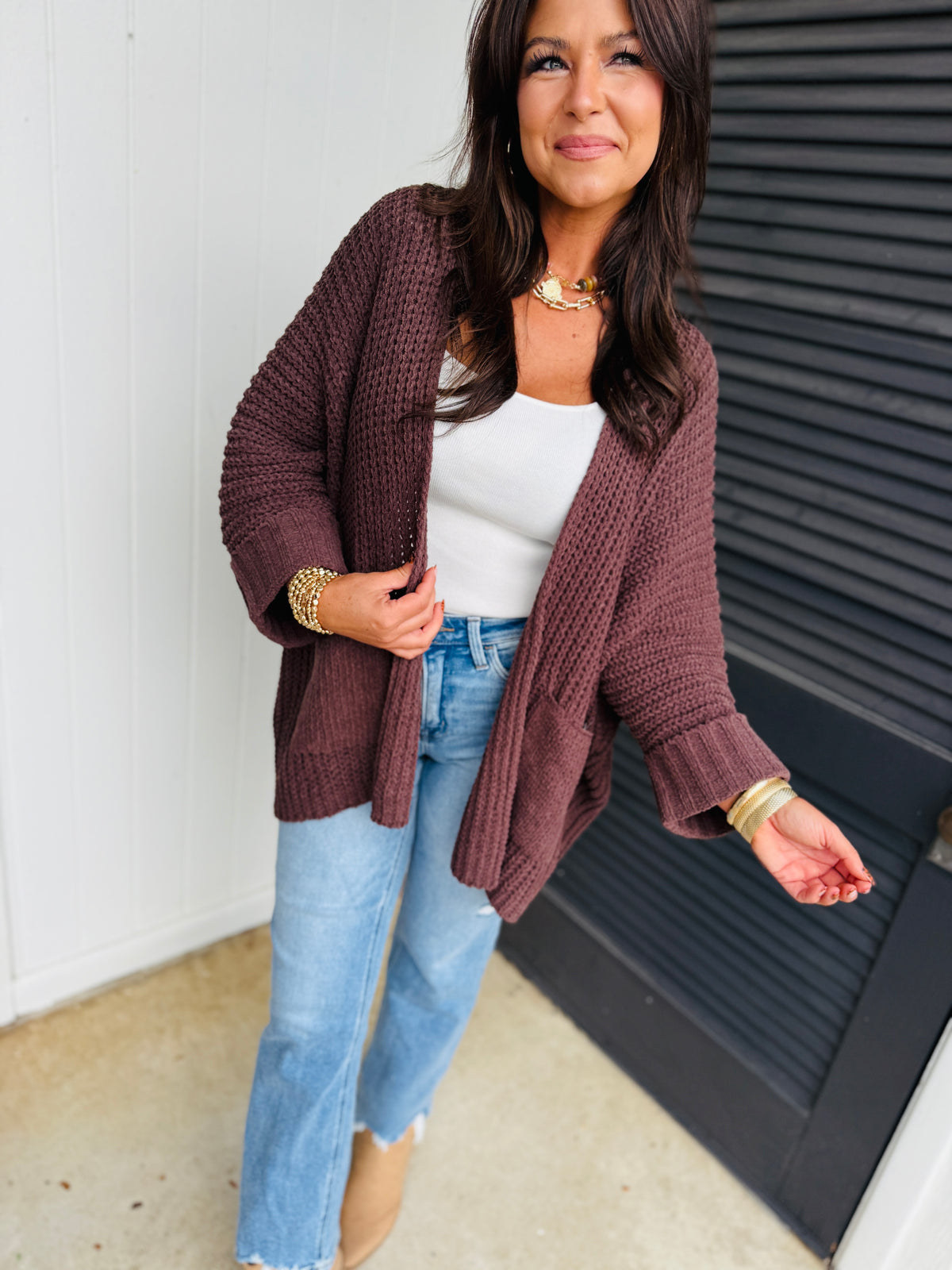 Woman wearing burgundy knit cardigan over white top with blue jeans and gold jewelry accessories.
