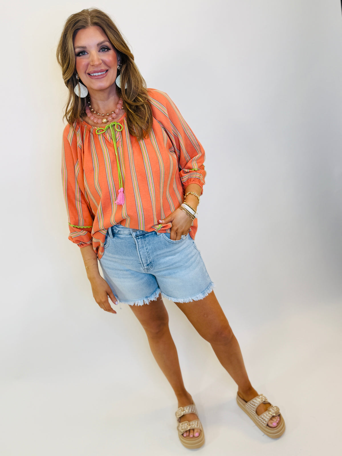 Woman wearing orange striped peasant blouse with tie neckline, denim shorts, and layered jewelry accessories.