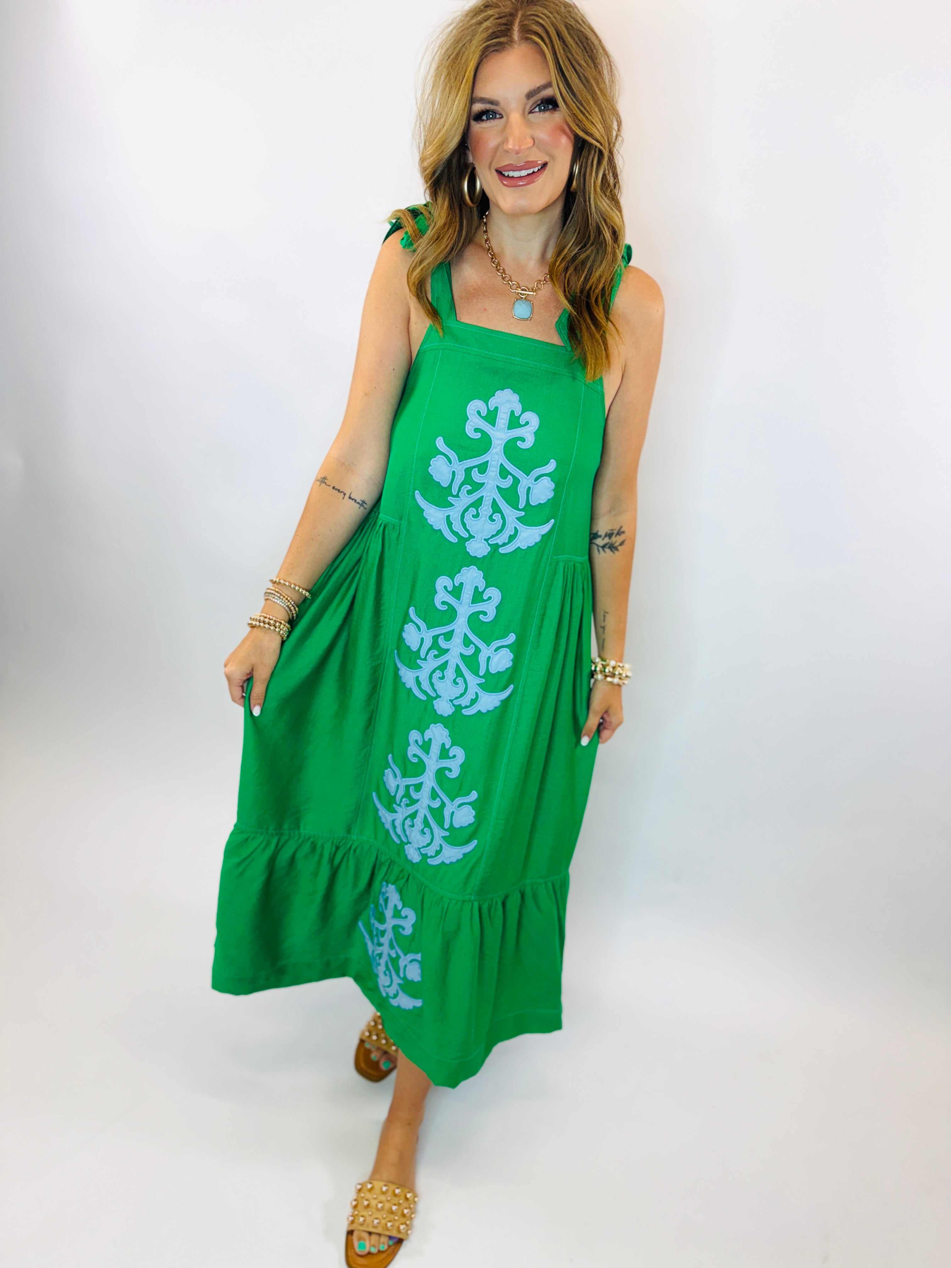 Woman wearing green maxi dress with blue embroidered floral pattern, tie shoulder straps, and tiered ruffle hem.
