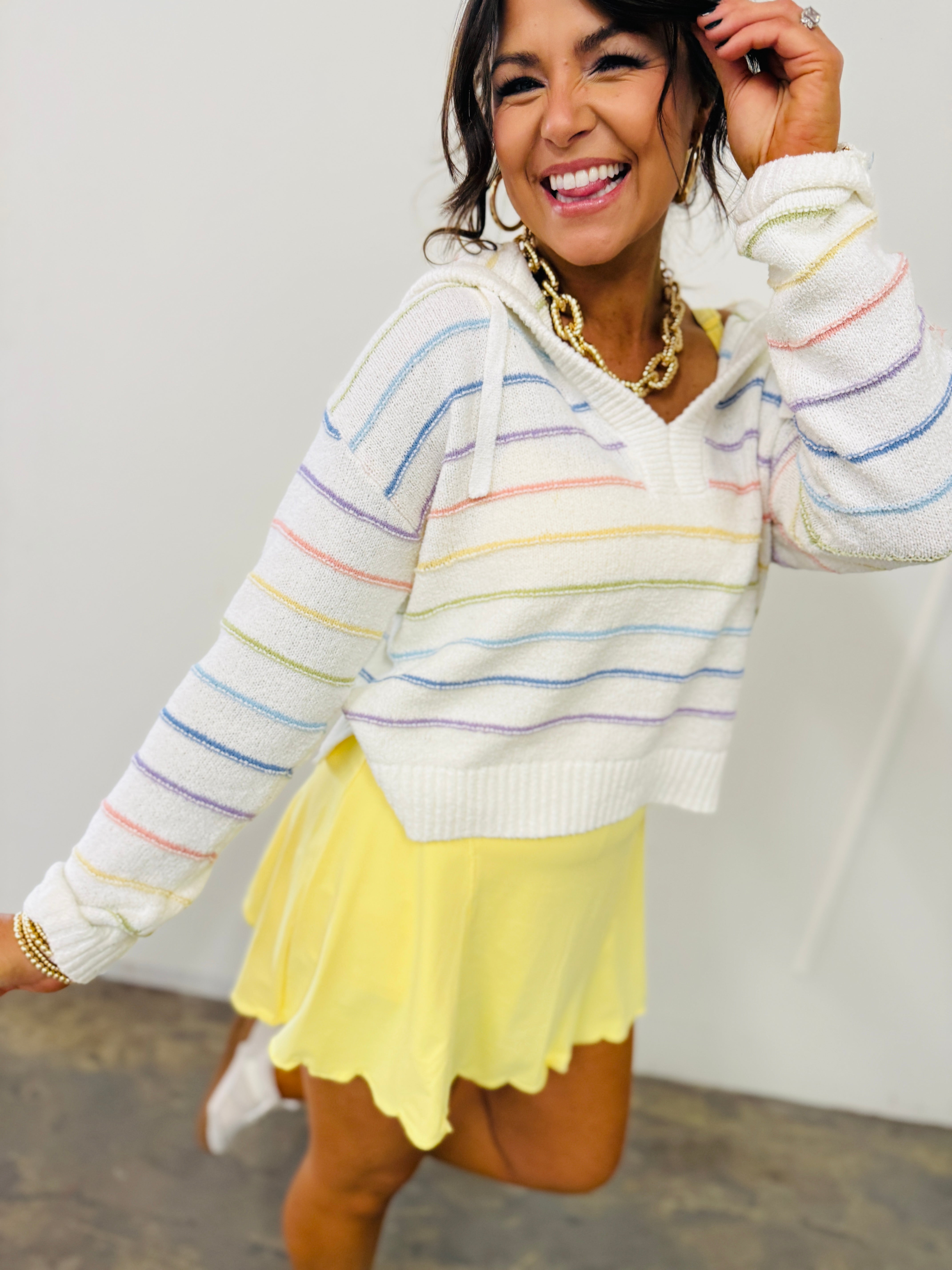 Woman wearing white striped hooded sweater with colorful rainbow stripes and yellow pleated mini skirt, smiling cheerfully.