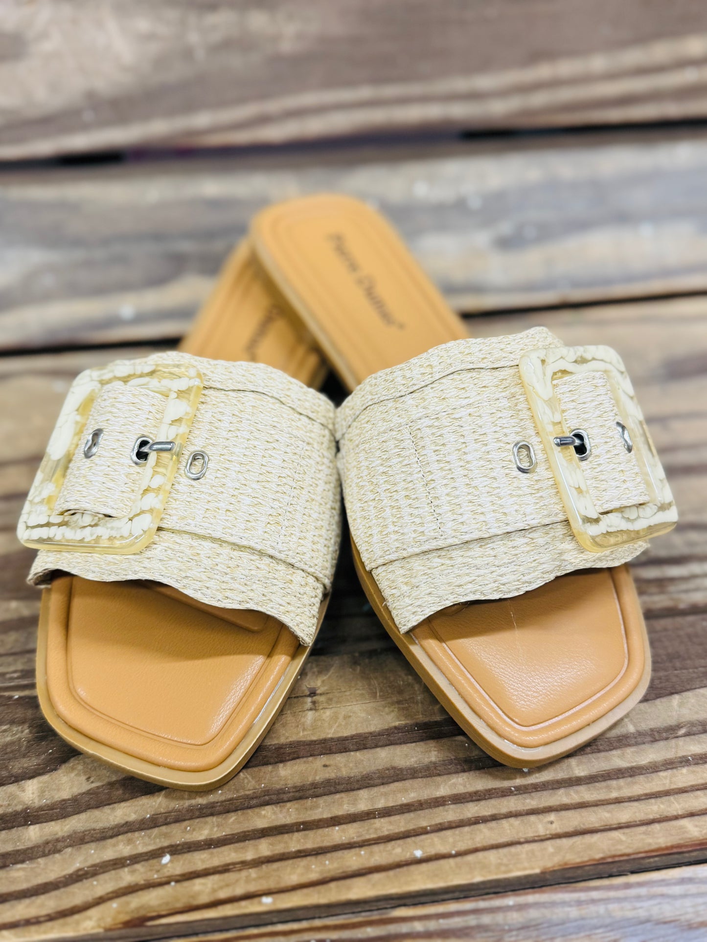 Beige textured slide sandals with buckle straps and square toes on wooden deck surface
