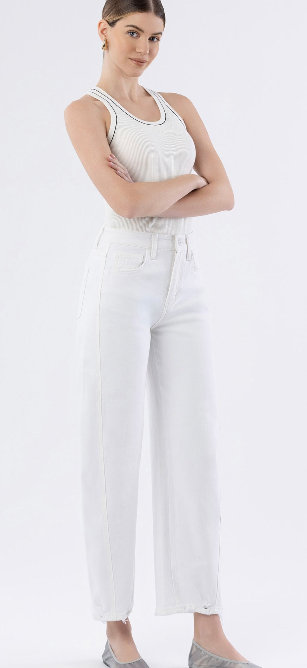 Woman wearing white ribbed tank top and high-waisted white barrel jeans, arms crossed, against white background.