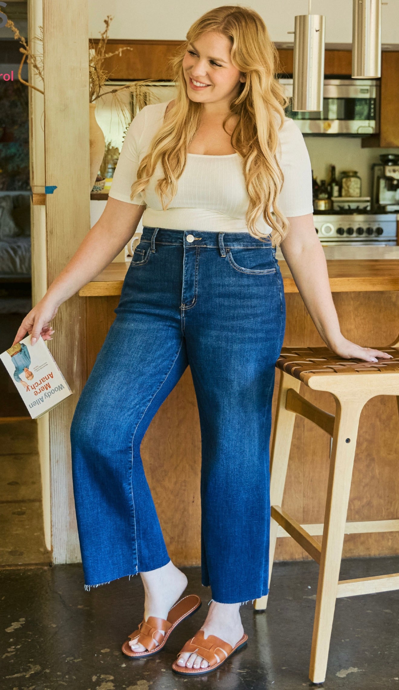 Woman wearing blue wide leg cropped jeans with white top and brown sandals in modern kitchen