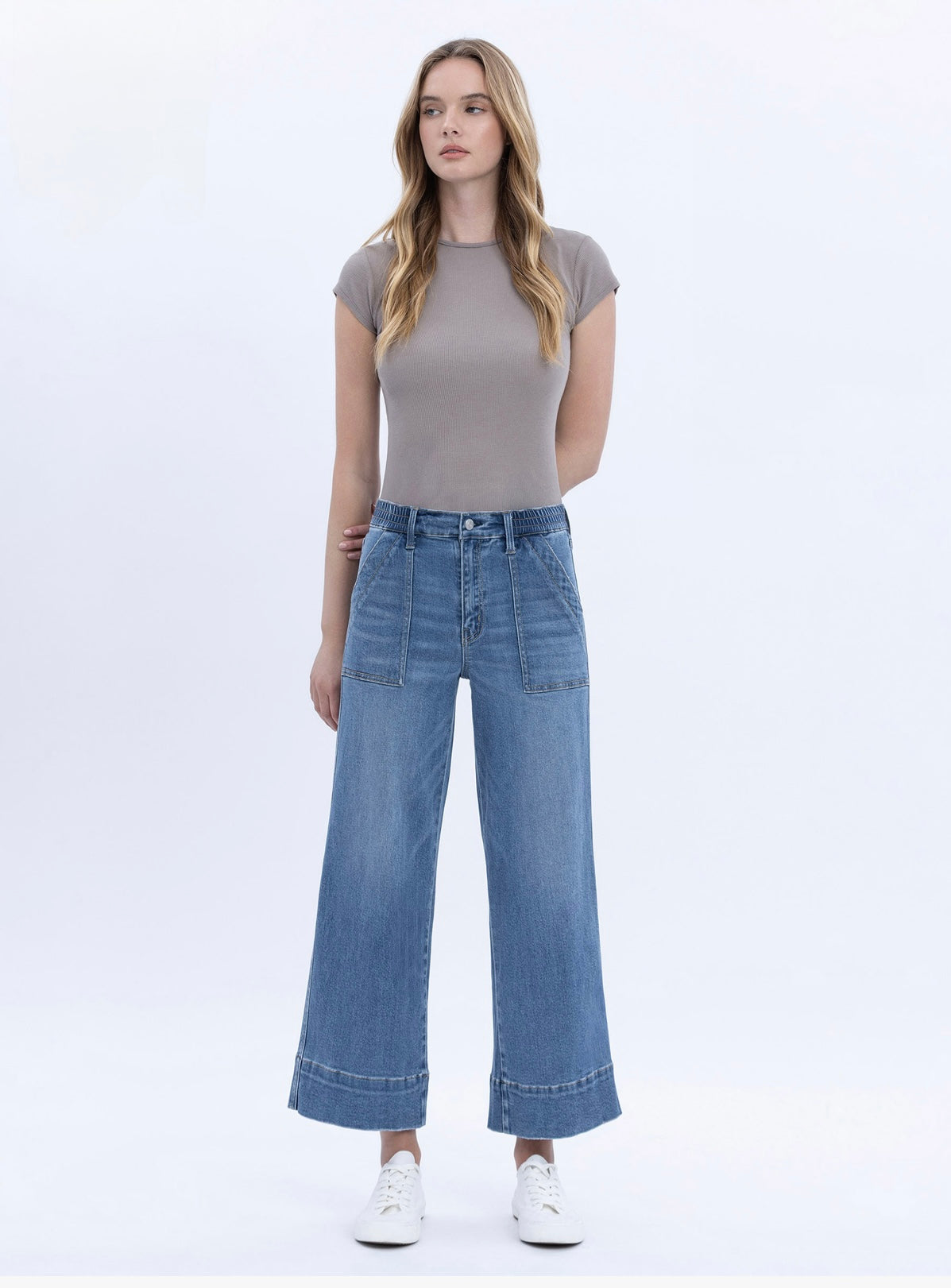 Woman wearing gray fitted t-shirt and blue wide-leg elastic waist denim pants with white sneakers on white background.