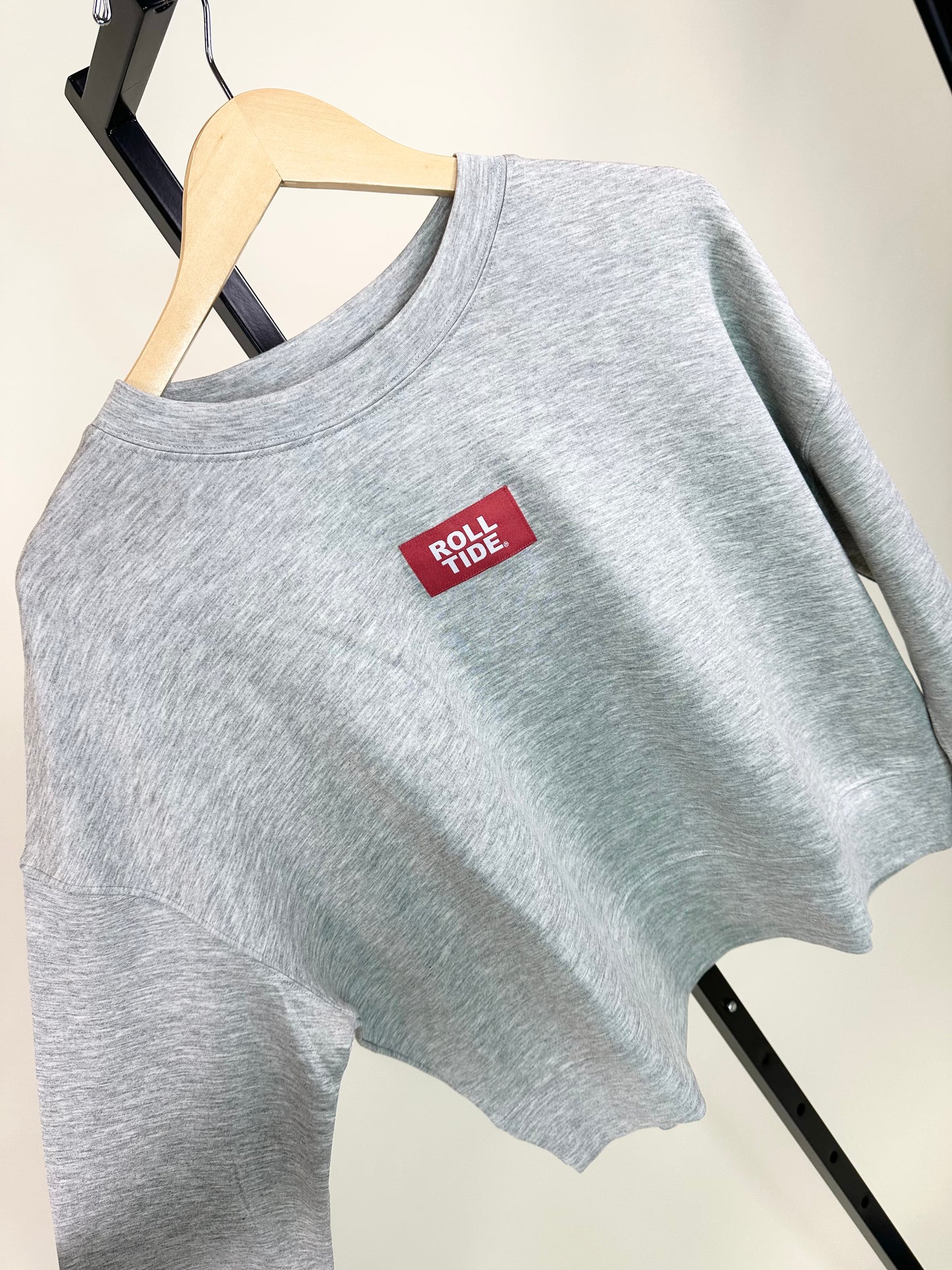 Gray heather crewneck sweatshirt with small crimson "Roll Tide" woven label on chest, displayed on wooden hanger.