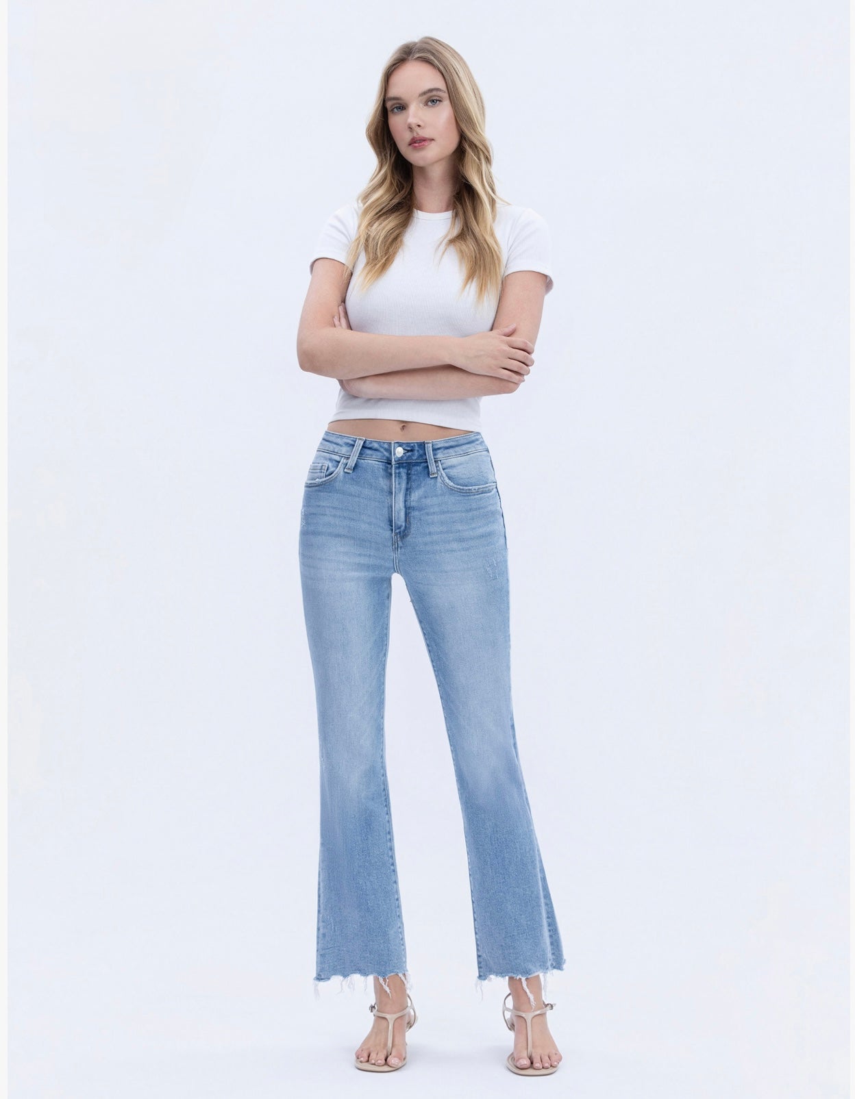 Woman wearing light blue flare jeans with frayed hem and white crop top, standing with arms crossed.