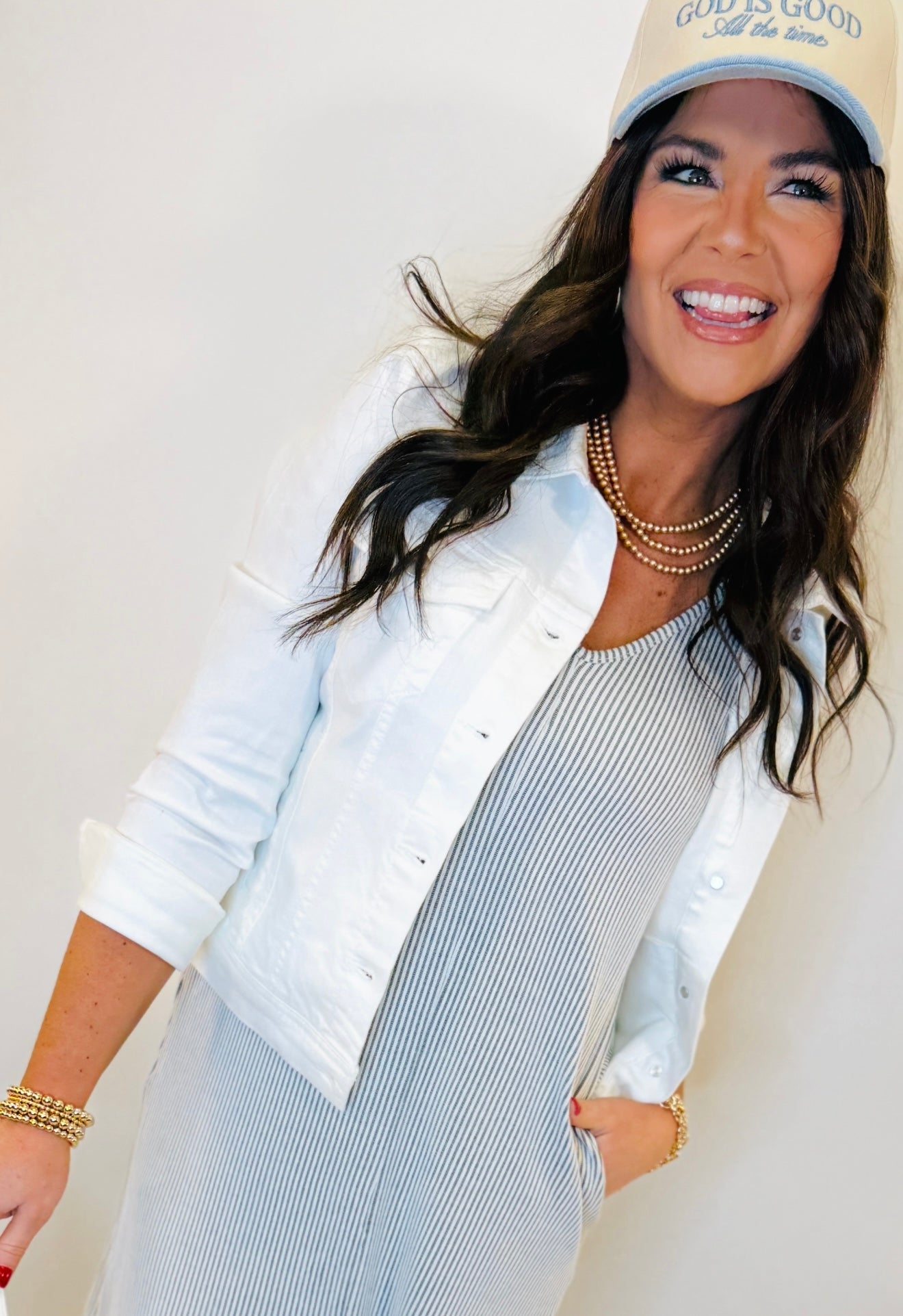 Woman wearing white denim jacket over blue striped dress with pearl necklace and baseball cap, smiling