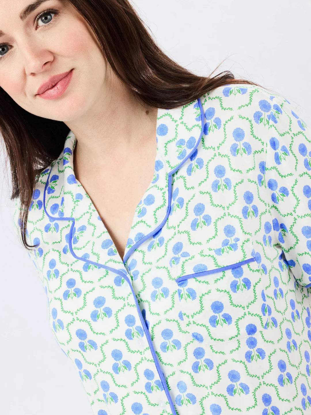 Woman wearing white pajama top with blue floral pattern and green vine details, featuring blue piped trim.