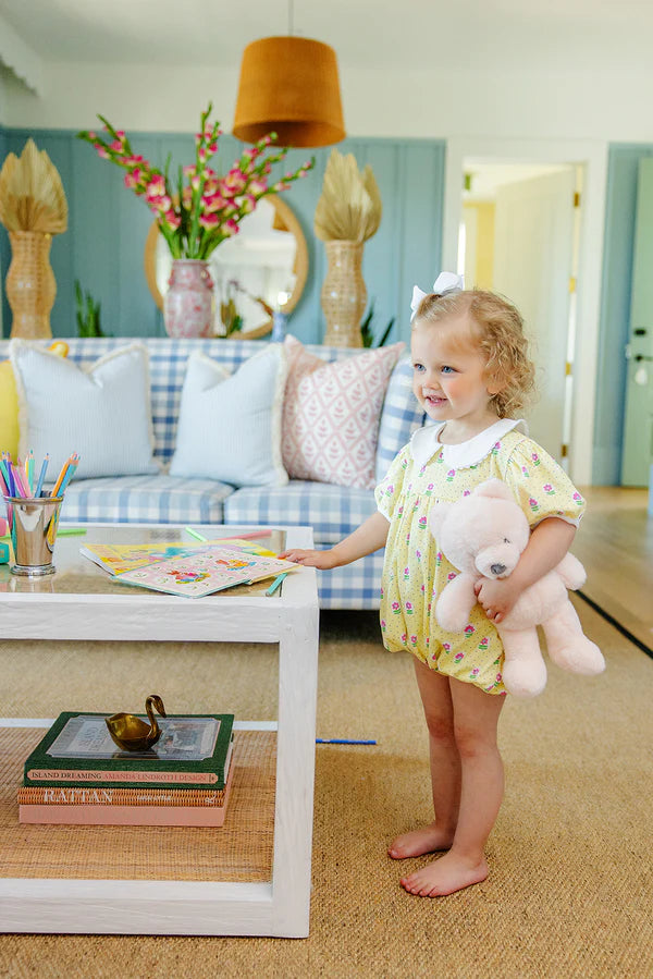 Toddler wearing yellow floral Pima cotton bubble romper while holding pink stuffed animal in bright living room