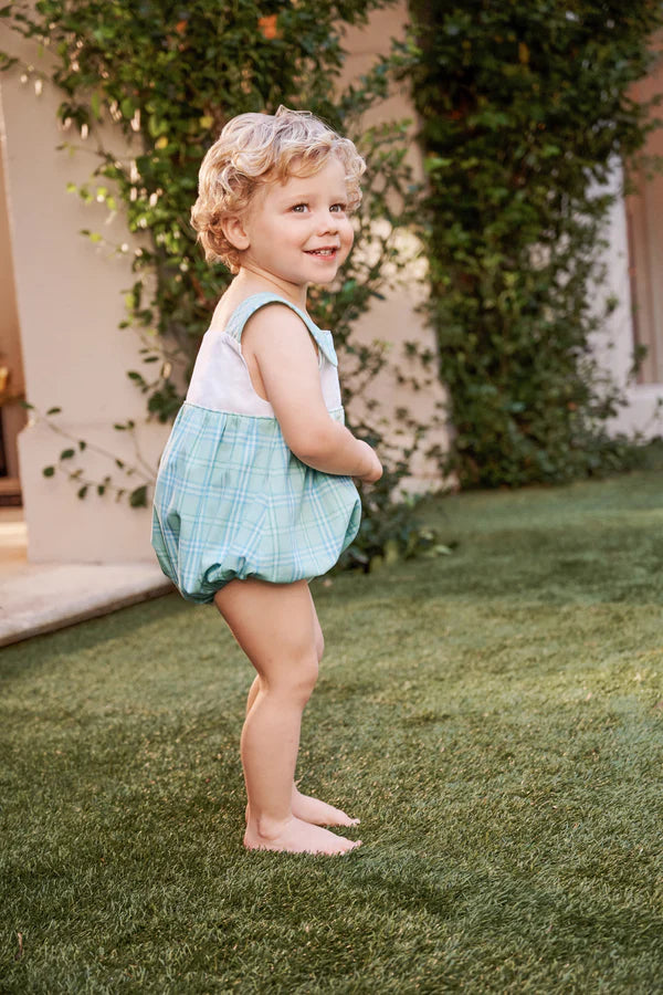 Smiling toddler wearing light blue plaid bubble romper standing on grass in garden setting