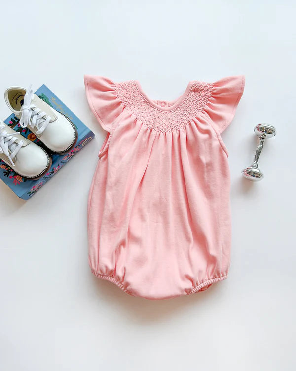 Pink baby romper with smocked neckline and flutter sleeves displayed with white shoes and rattle