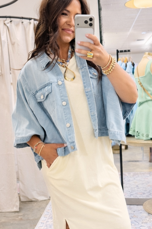 Woman wearing cream-colored Deja t-shirt maxi dress with oversized denim jacket, taking mirror selfie in retail store.