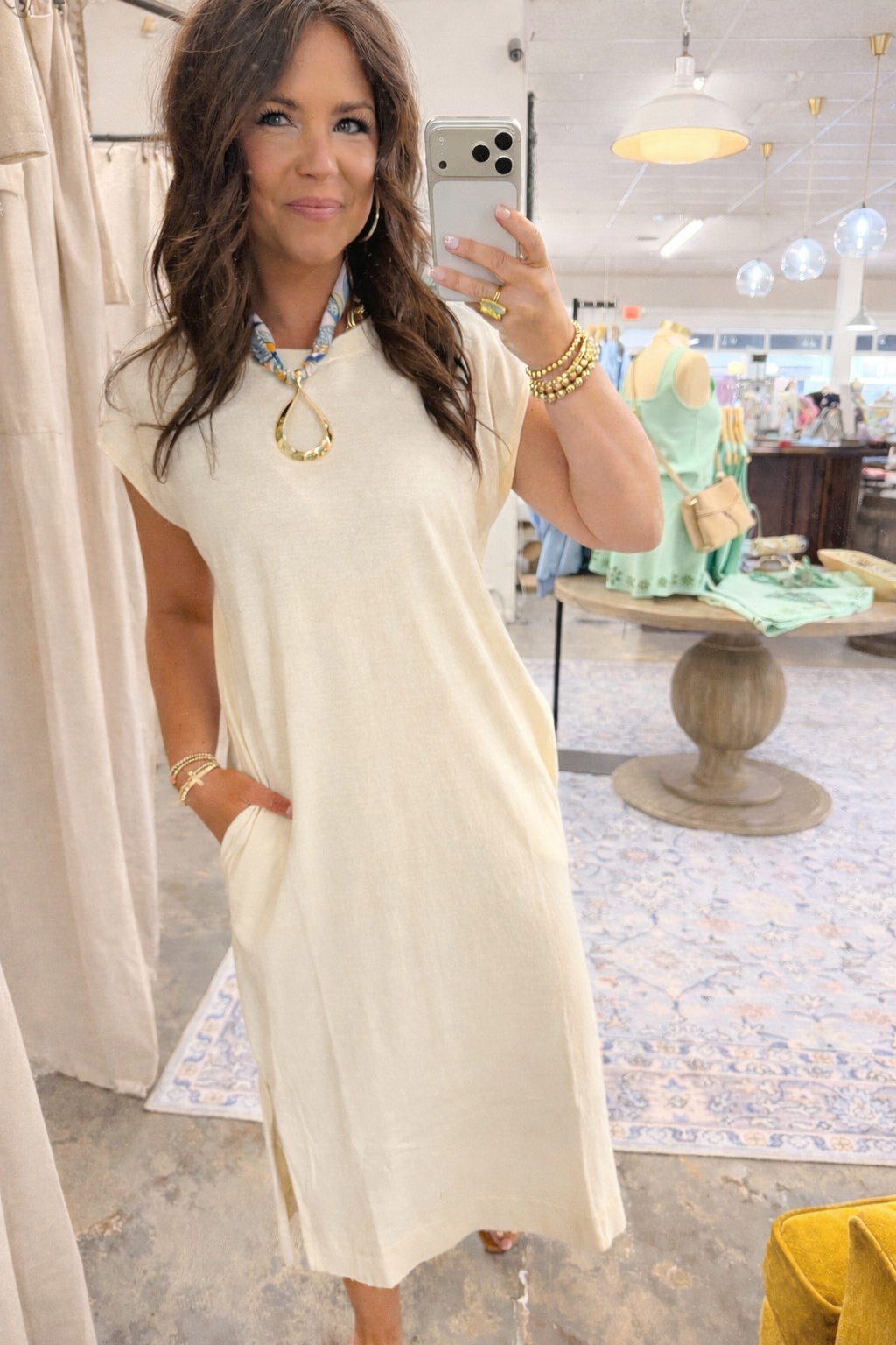 Woman wearing cream-colored Deja t-shirt maxi dress with gold jewelry, taking mirror selfie in boutique setting.