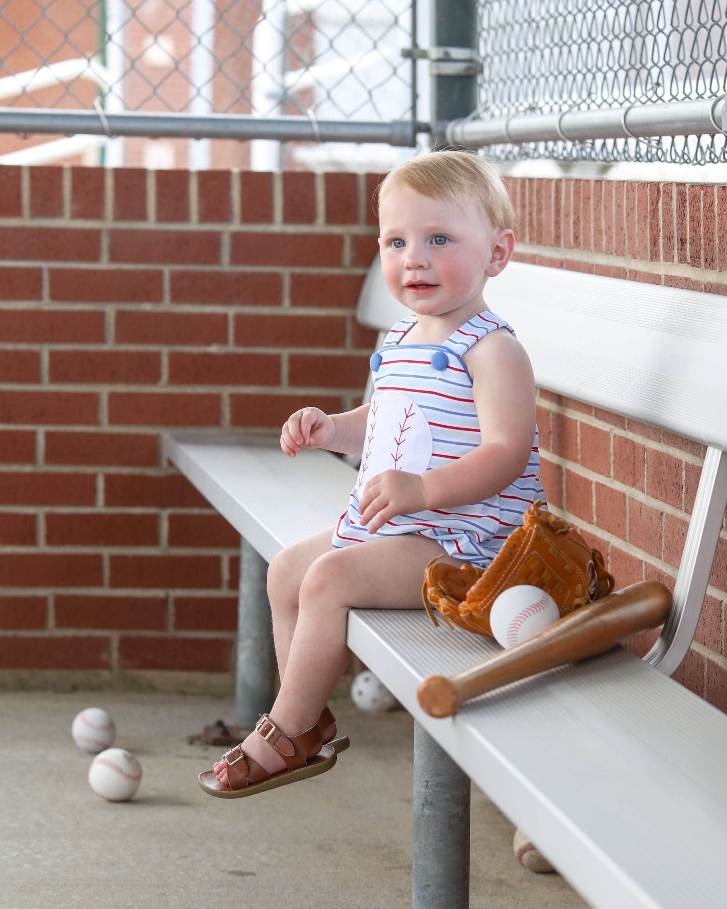 Young child sitting on bleacher bench wearing striped baseball-themed sunsuit with baseball glove and balls nearby.