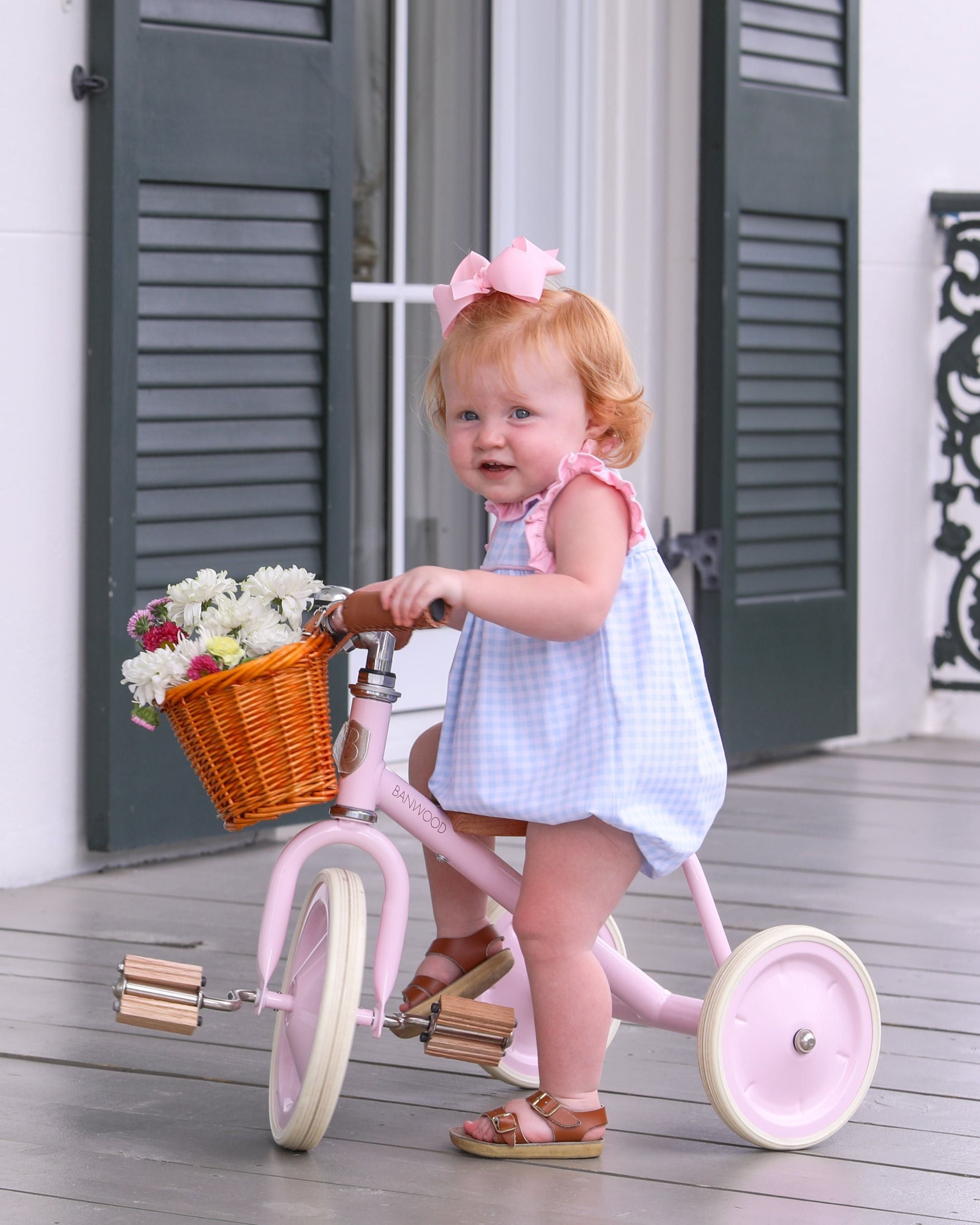 Young child wearing white bubble romper with pink bow, standing beside pink tricycle with flower basket.