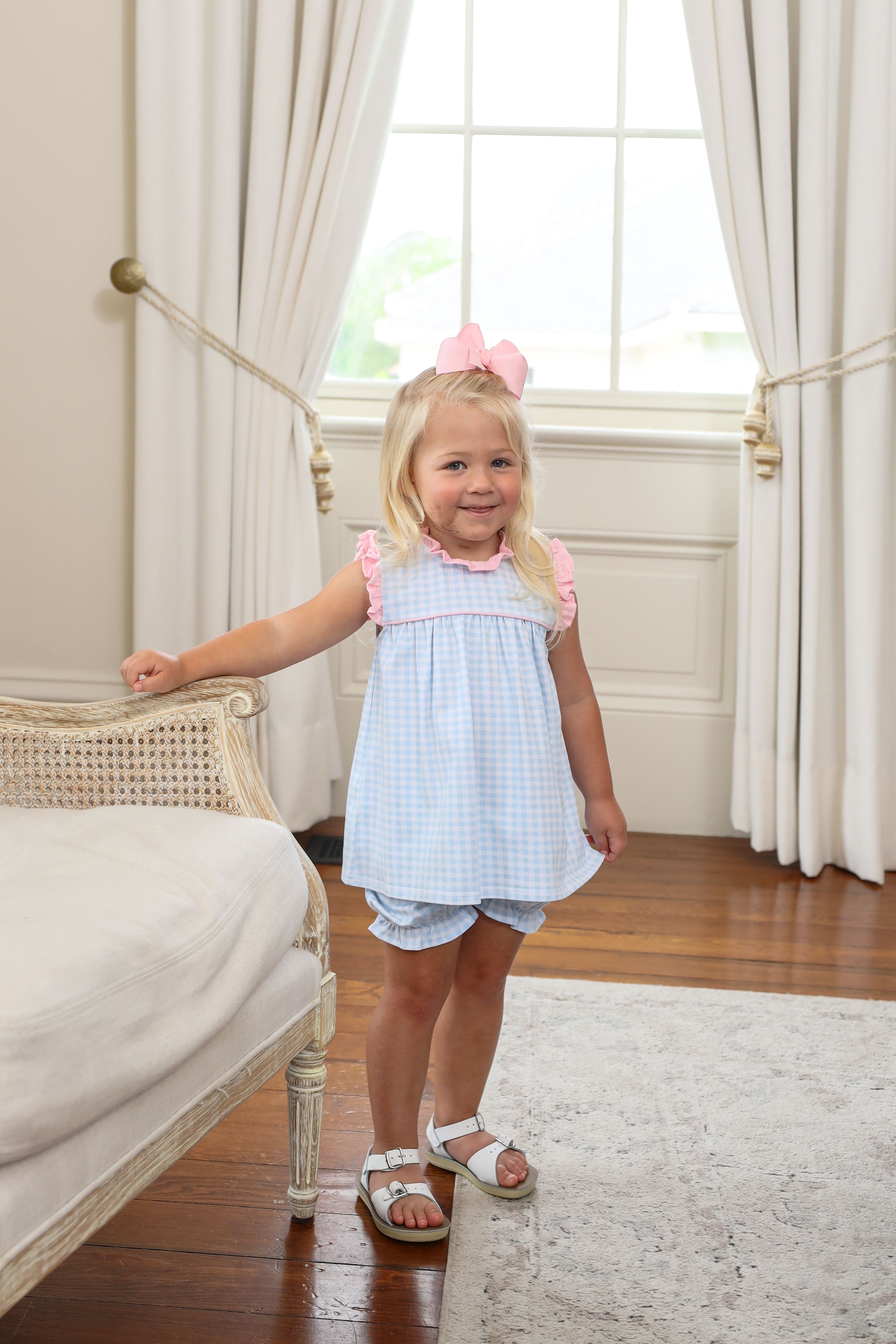 Young blonde girl wearing light blue gingham dress with pink trim and matching bloomers, standing beside vintage chair