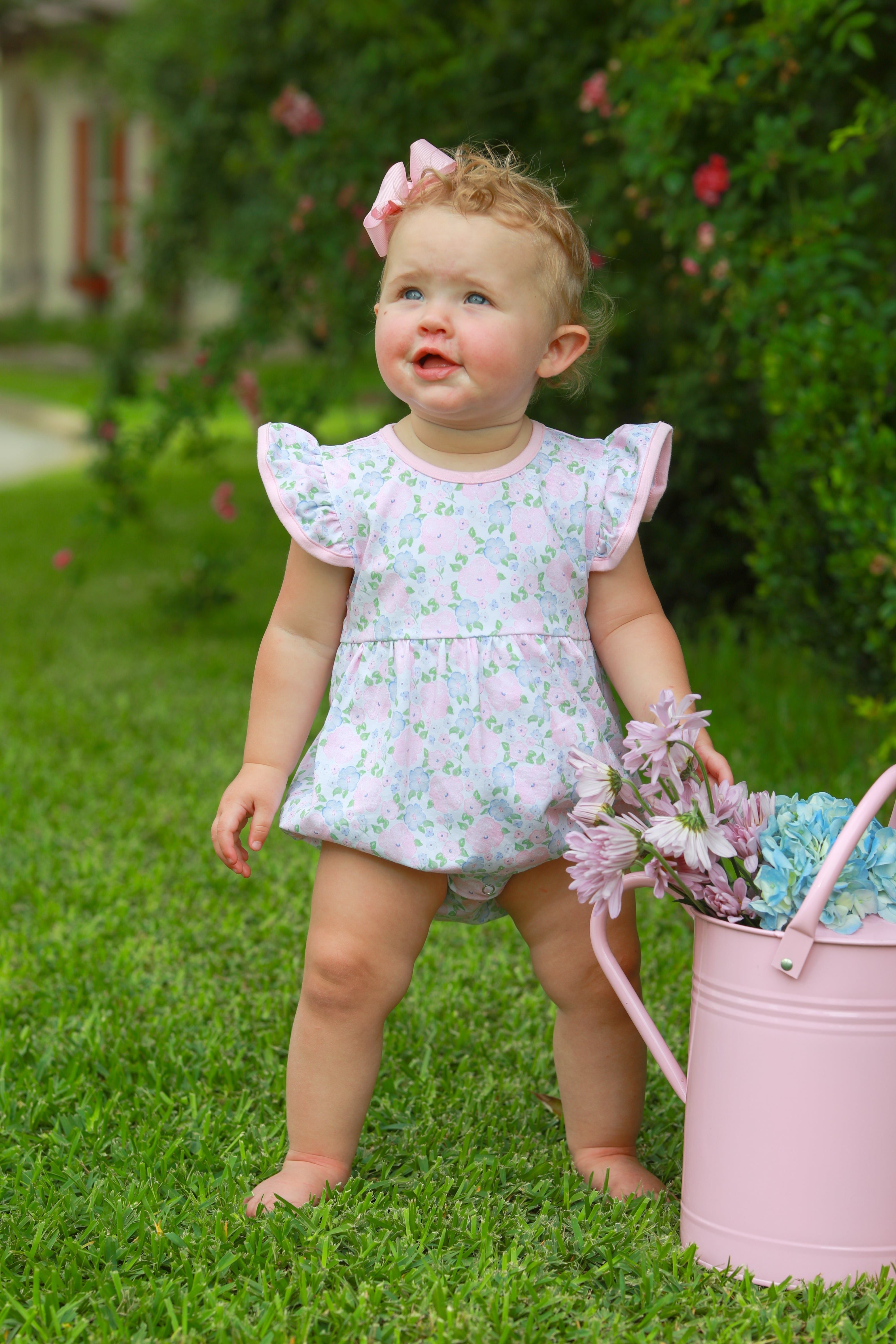 Baby wearing pink floral bubble romper with flutter sleeves, standing on grass holding pink bucket with flowers.